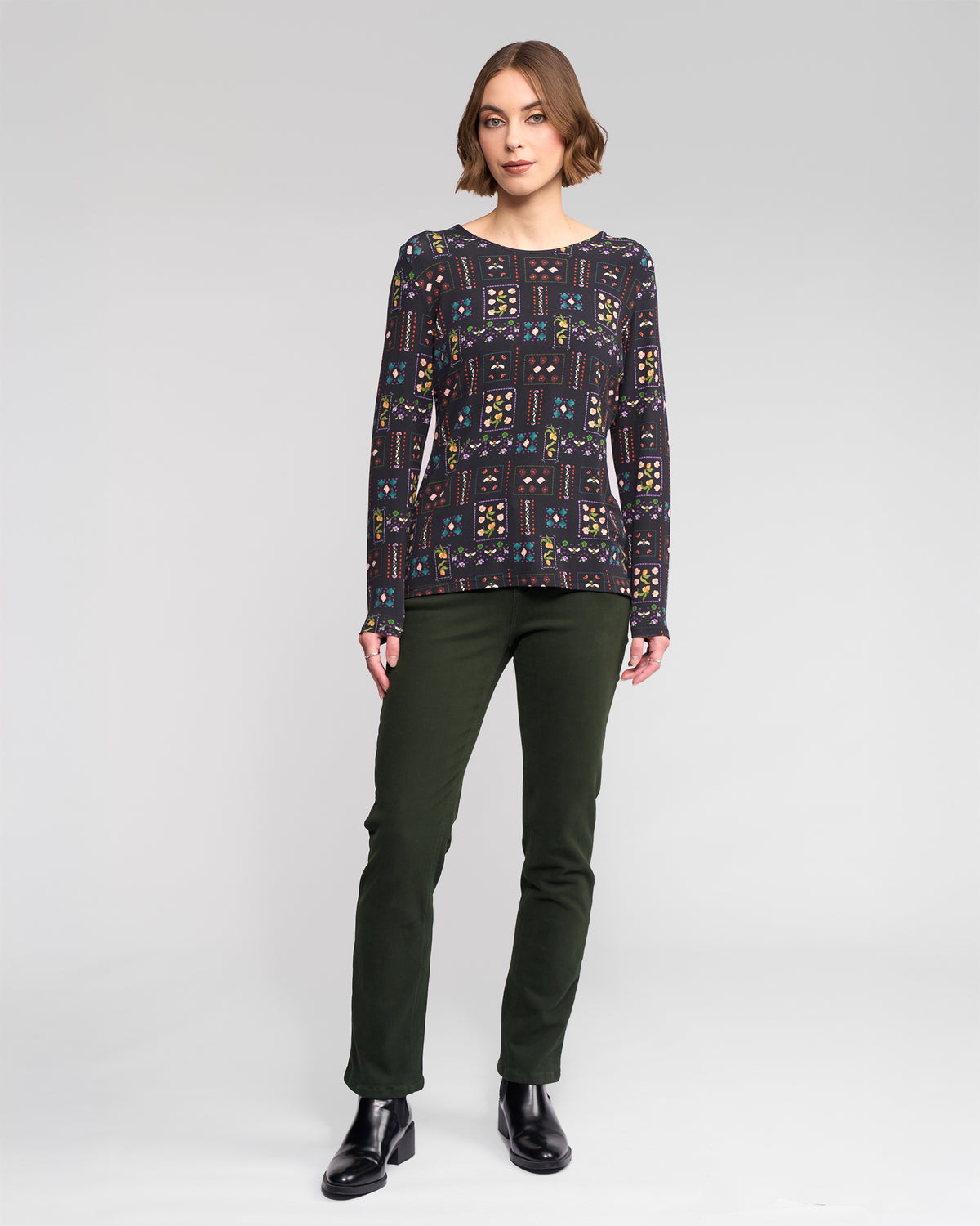 A woman with short brown hair stands against a light gray background, wearing the Vassalli Staple Top - Haven (a dark patterned long-sleeve stretch jersey), slim-fit dark green pants, and black ankle boots, looking forward with a neutral expression.