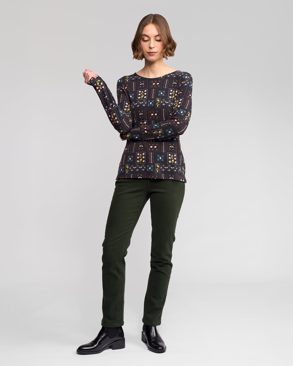 A person stands against a plain light background, wearing the Vassalli Staple Top - Haven (a slim-fit stretch jersey long-sleeve), dark green pants, and black ankle boots, with one arm bent and the other relaxed.
