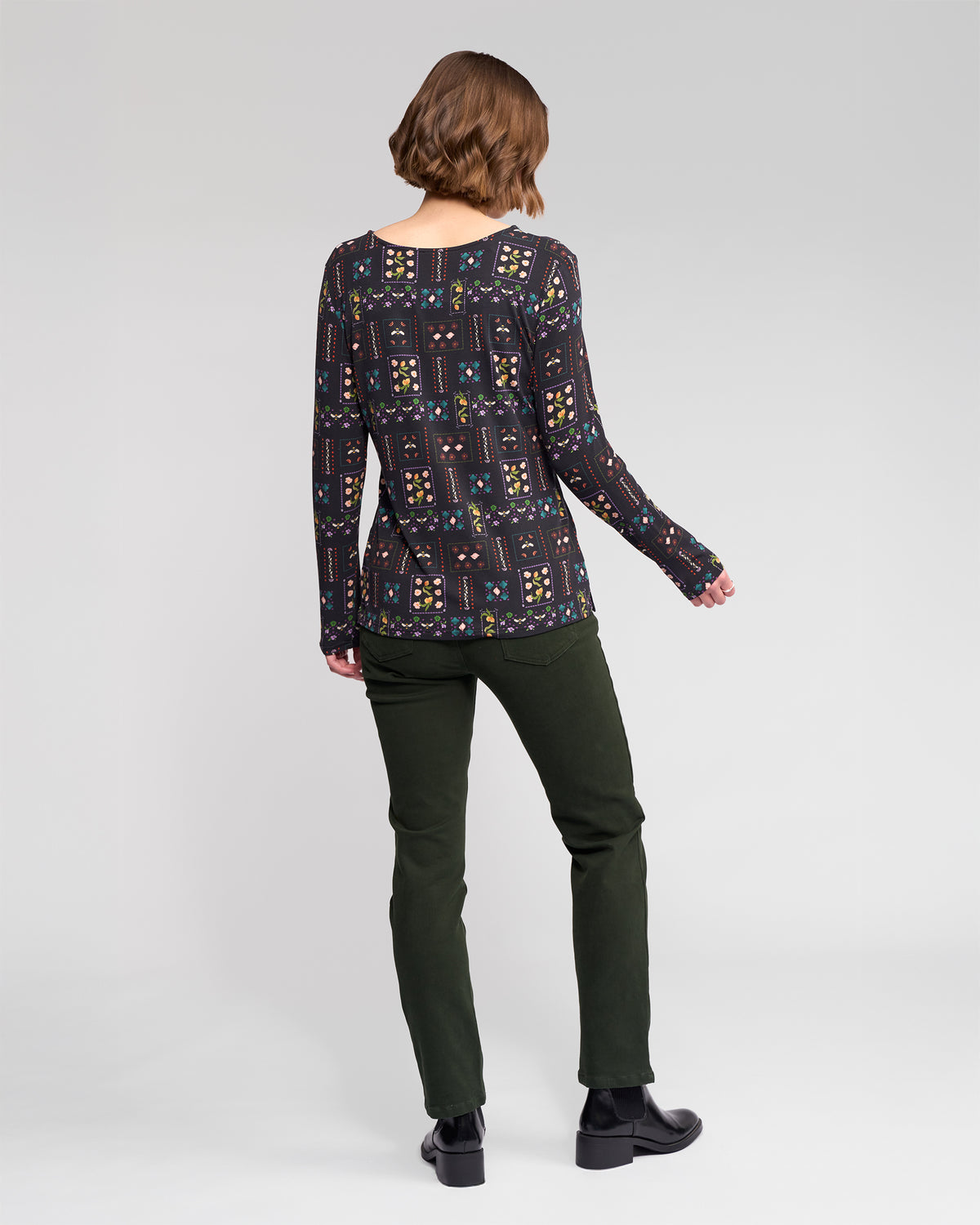 A person with short brown hair, wearing the Vassalli Staple Top - Haven, dark green pants, and black shoes stands facing away from the camera against a plain light grey background.