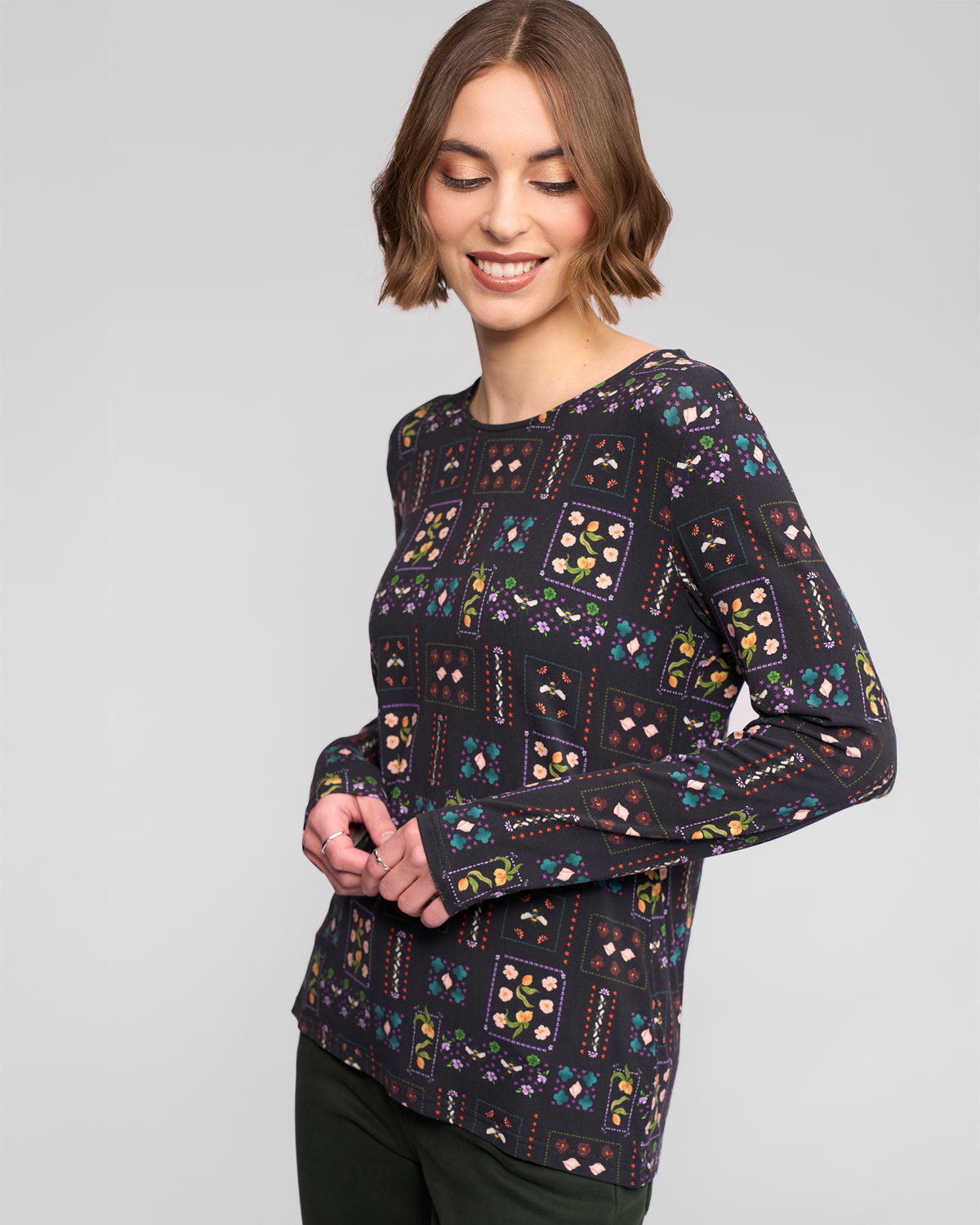 A woman with short brown hair smiles while looking down, wearing the Vassalli Staple Top - Haven, a slim-fit long-sleeve with a colorful geometric and floral pattern, paired with dark pants against a plain light background.
