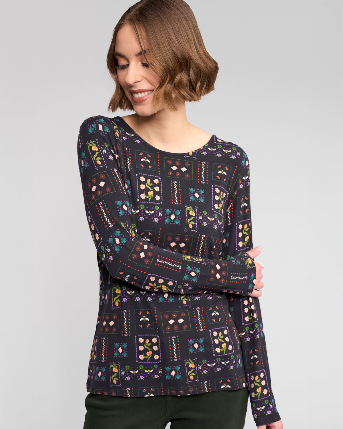 A woman with short brown hair smiles while wearing the Vassalli Staple Top - Haven, a slim-fit long-sleeve top with a colorful geometric and floral pattern. She stands against a plain light background, crossing her arms and looking down to the side.