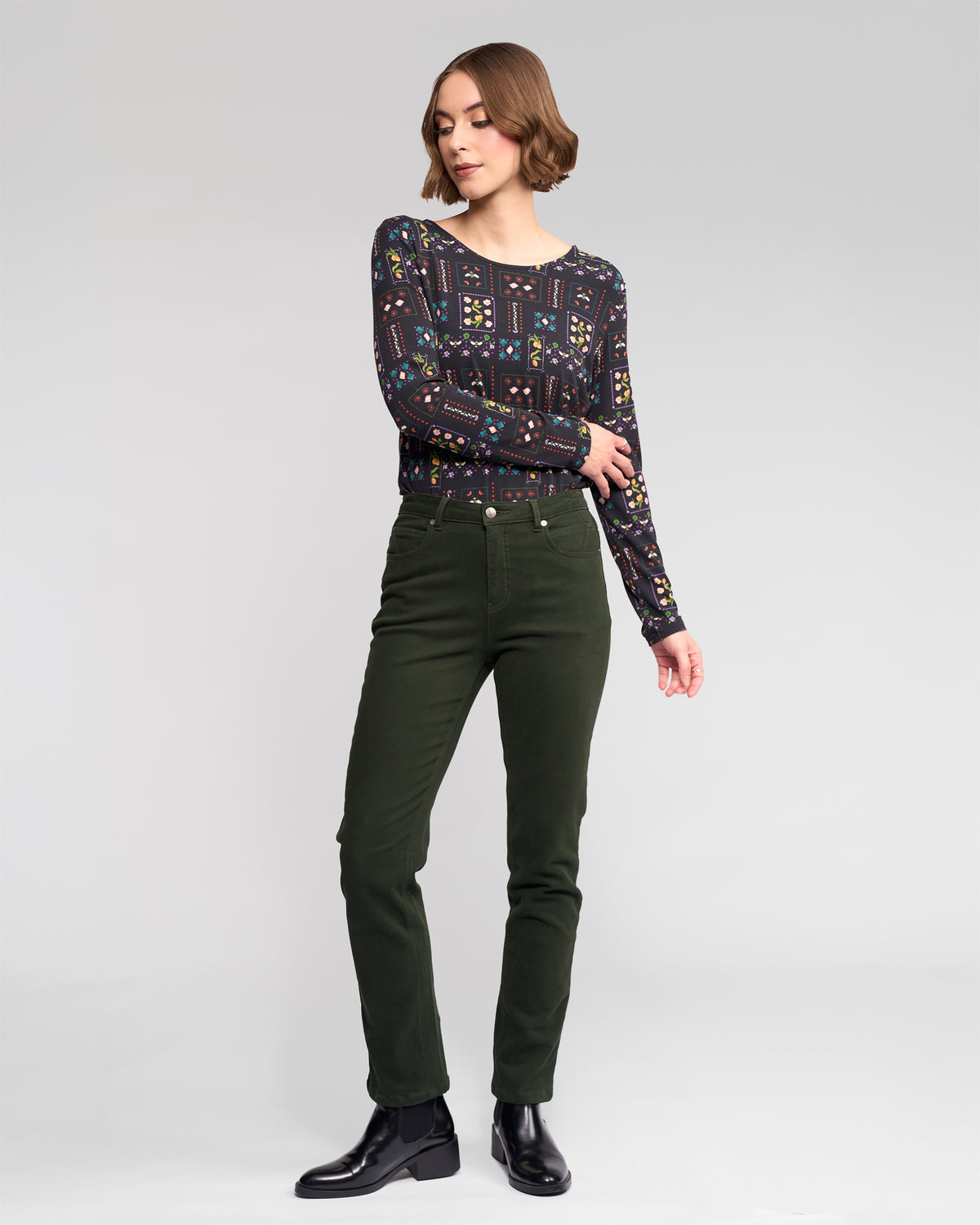 A woman with short brown hair wears the Vassalli Staple Top - Haven, paired with slim-fit dark green pants and black ankle boots. She stands against a plain light gray background, looking to her left with one hand slightly raised.