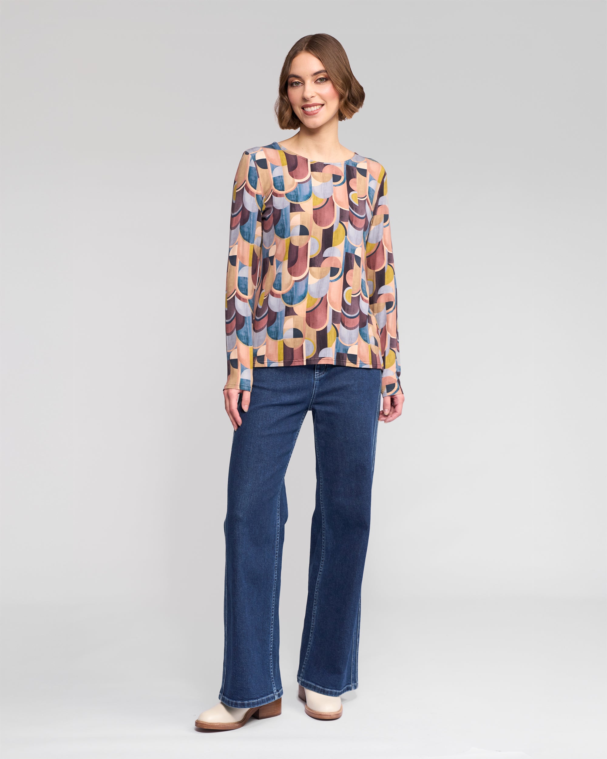 A woman smiles, wearing the Vassalli Staple Top - Revolve with a colorful retro print in stretch jersey, paired with blue flared jeans and cream shoes, standing against a plain light gray background.