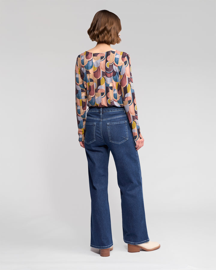 A person with short brown hair faces away, wearing the Vassalli Staple Top - Revolve, featuring a colorful retro print and long sleeves, paired with high-waisted wide-leg blue jeans and beige-heeled shoes against a plain light background.