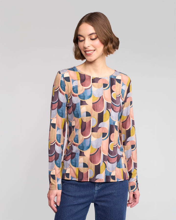 A woman with short brown hair smiles and looks down while wearing the Vassalli Staple Top - Revolve, a retro stretch jersey long-sleeve featuring colorful geometric circles and curves, paired with blue jeans.