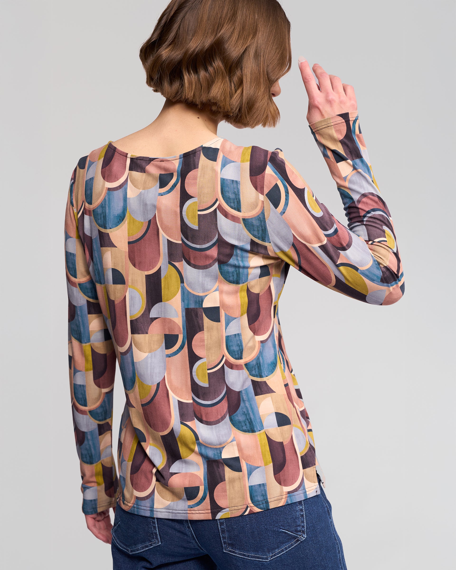 A person with short brown hair, seen from behind, wears the Vassalli Staple Top - Revolve: a long-sleeve retro top with a colorful geometric print in blue, yellow, brown, and pink, paired with blue jeans.