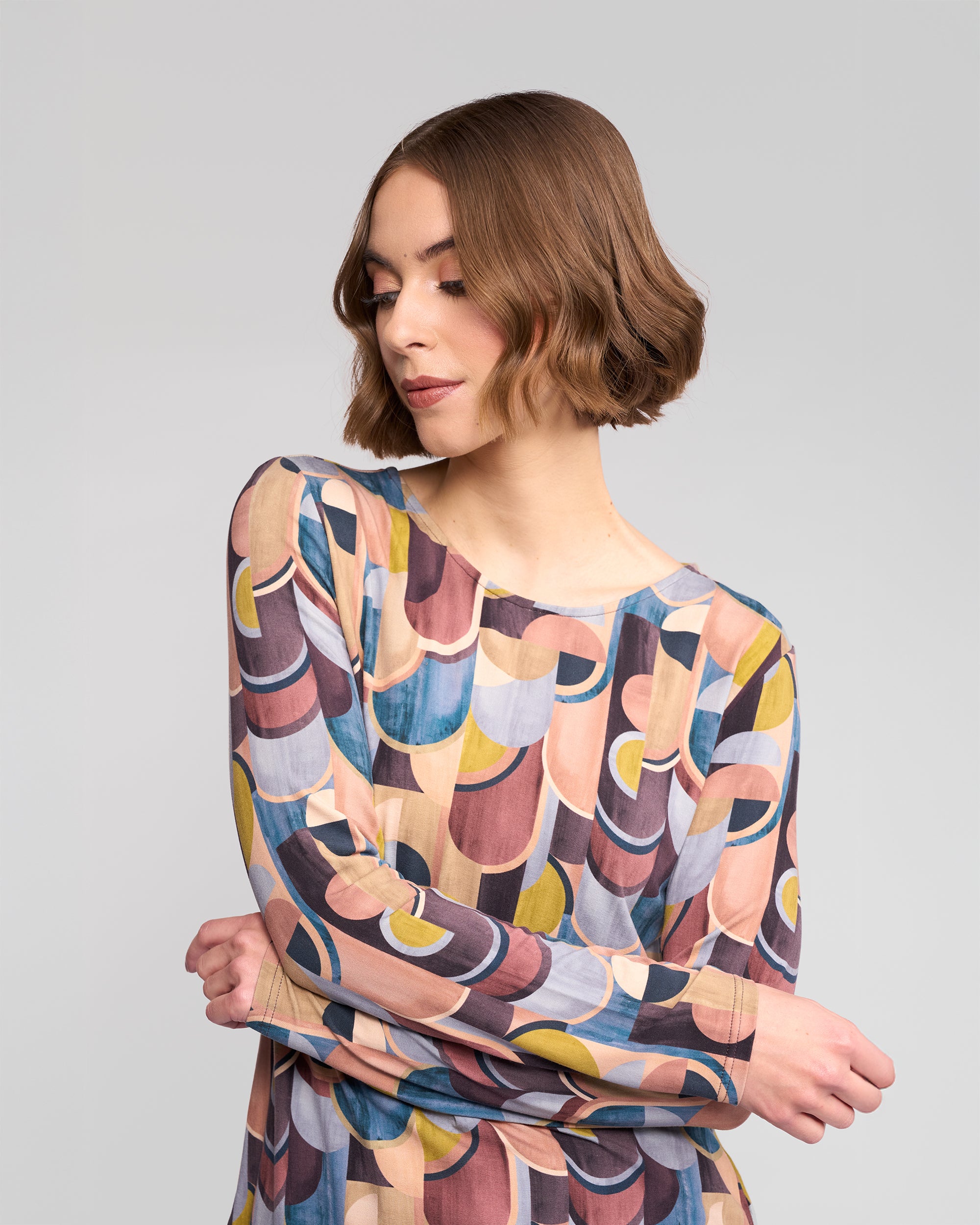 A woman with short brown hair wears the Vassalli Staple Top - Revolve, featuring a colorful geometric retro print, crossing her arms and looking down against a plain light gray background.