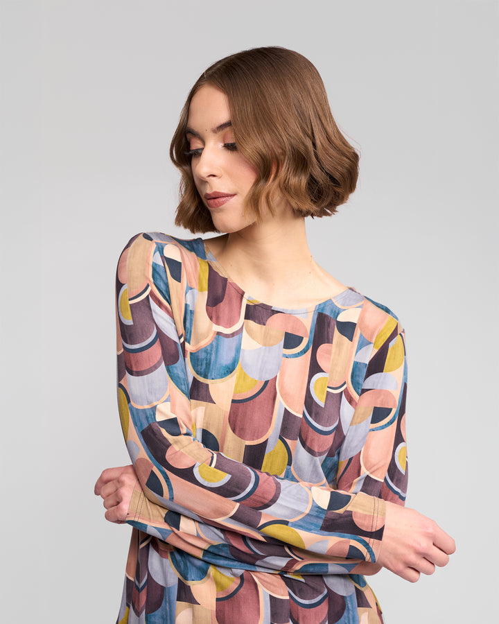 A woman with short brown hair wears the Vassalli Staple Top - Revolve, featuring a colorful geometric retro print, crossing her arms and looking down against a plain light gray background.