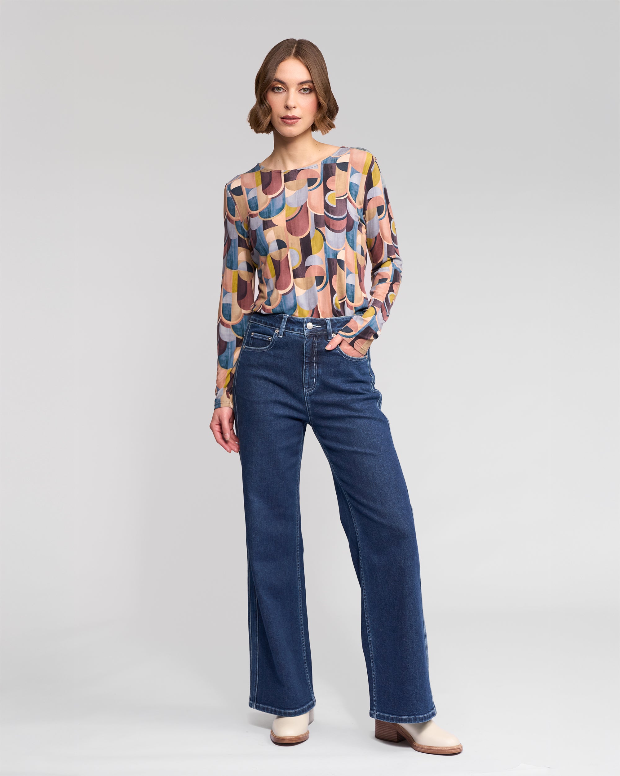 A woman with short wavy brown hair and a neutral expression wears the Vassalli Staple Top - Revolve, a colorful retro print stretch jersey long-sleeve top, paired with high-waisted blue wide-leg jeans and beige shoes against a plain background.