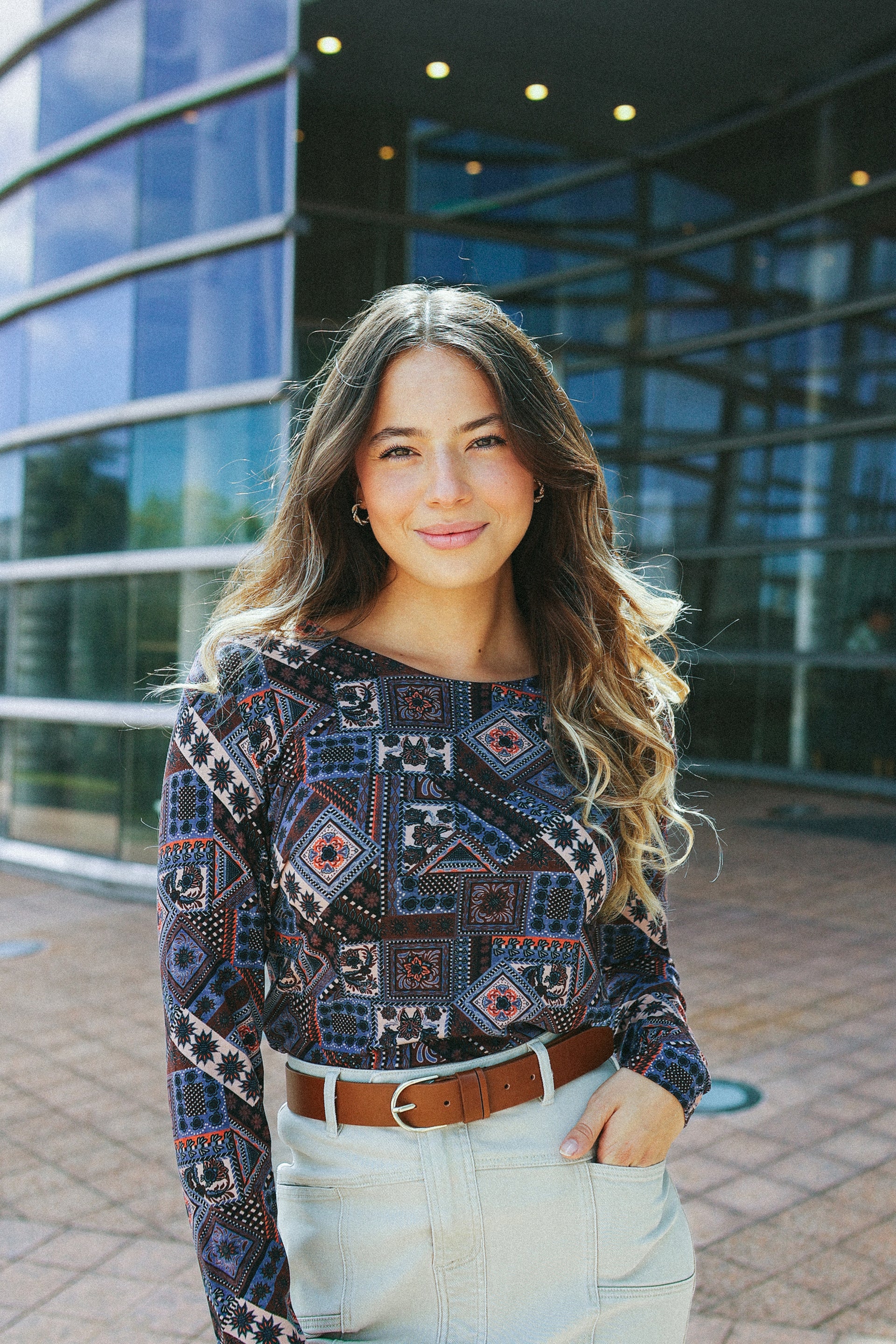 A young woman with long wavy hair, wearing the Vassalli Staple Top - Rue and a light skirt with a brown belt, stands confidently outside a modern glass building, smiling at the camera.