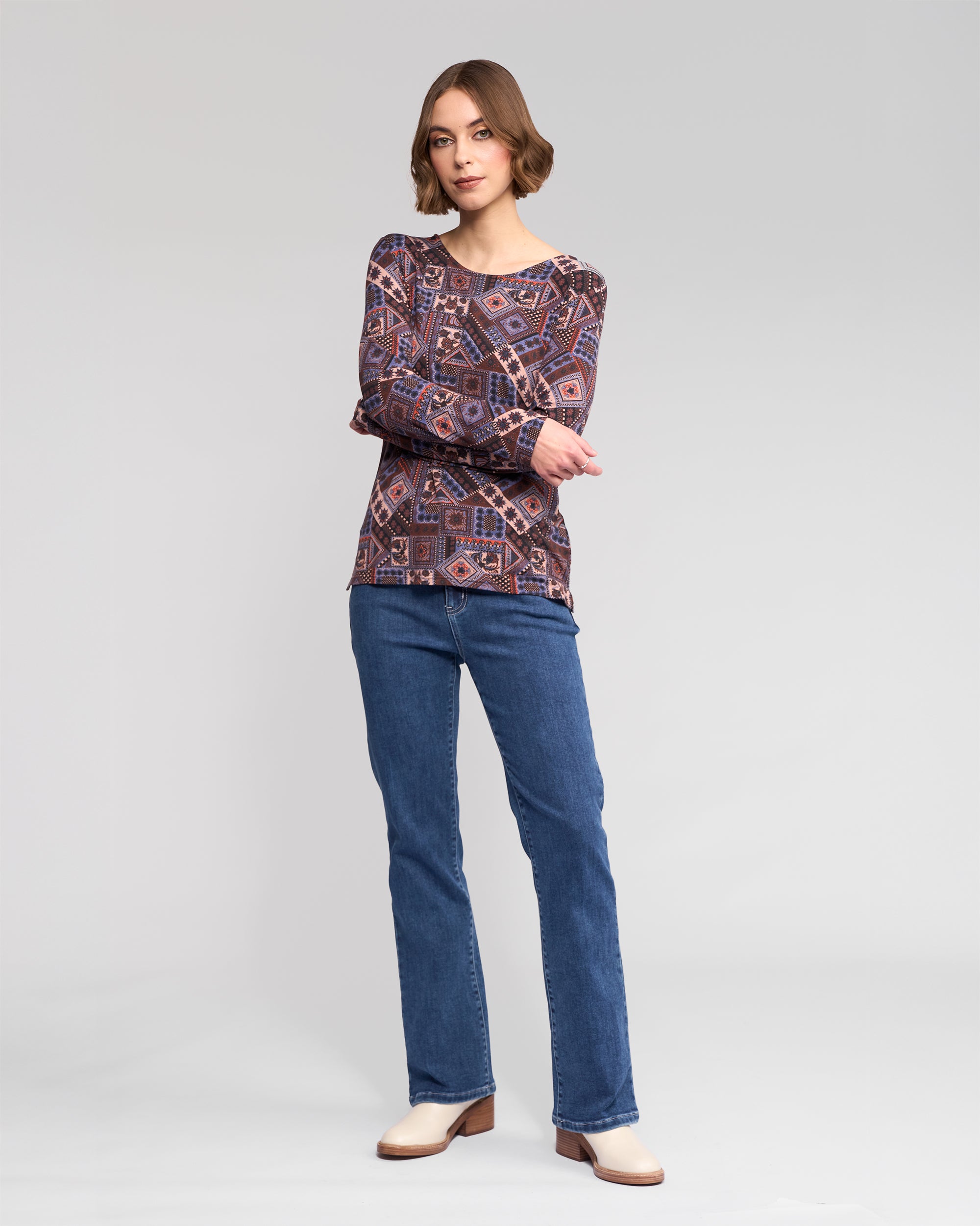 A woman with short, wavy hair stands against a light gray background, arms crossed, wearing the Vassalli Staple Top - Rue featuring geometric prints, paired with blue jeans and cream shoes.