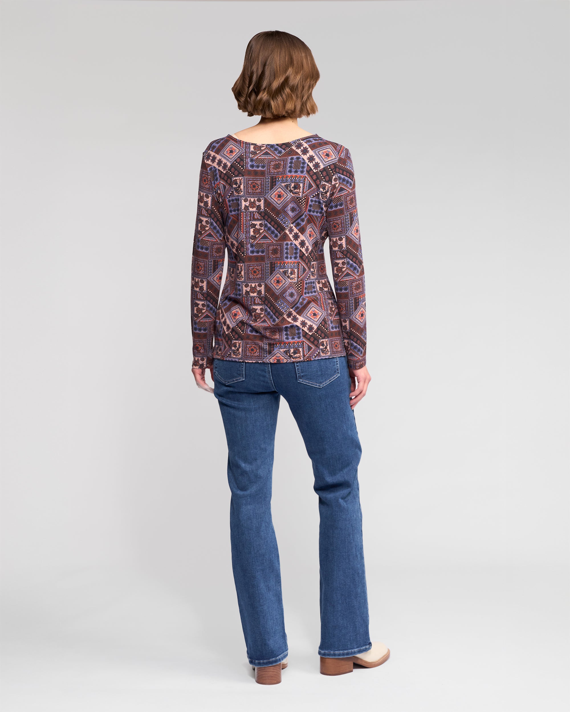 A person with short brown hair, wearing a Vassalli Staple Top - Rue in stretch jersey, blue jeans, and beige shoes stands facing away against a plain light gray background.