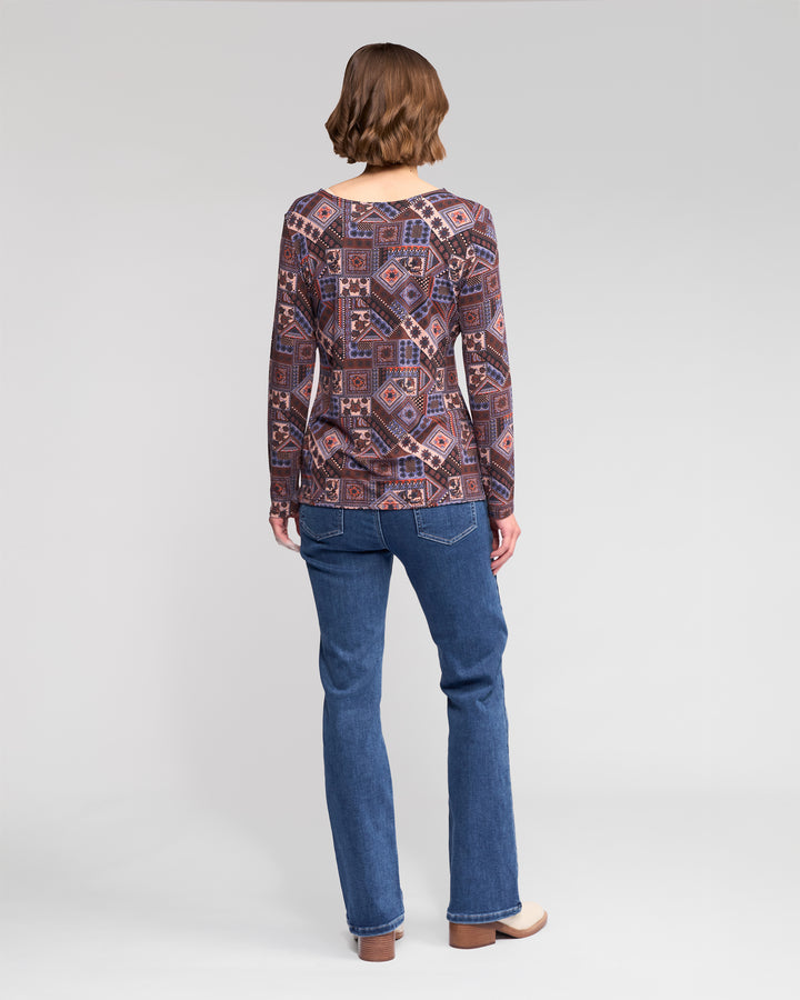 A person with short brown hair, wearing a Vassalli Staple Top - Rue in stretch jersey, blue jeans, and beige shoes stands facing away against a plain light gray background.