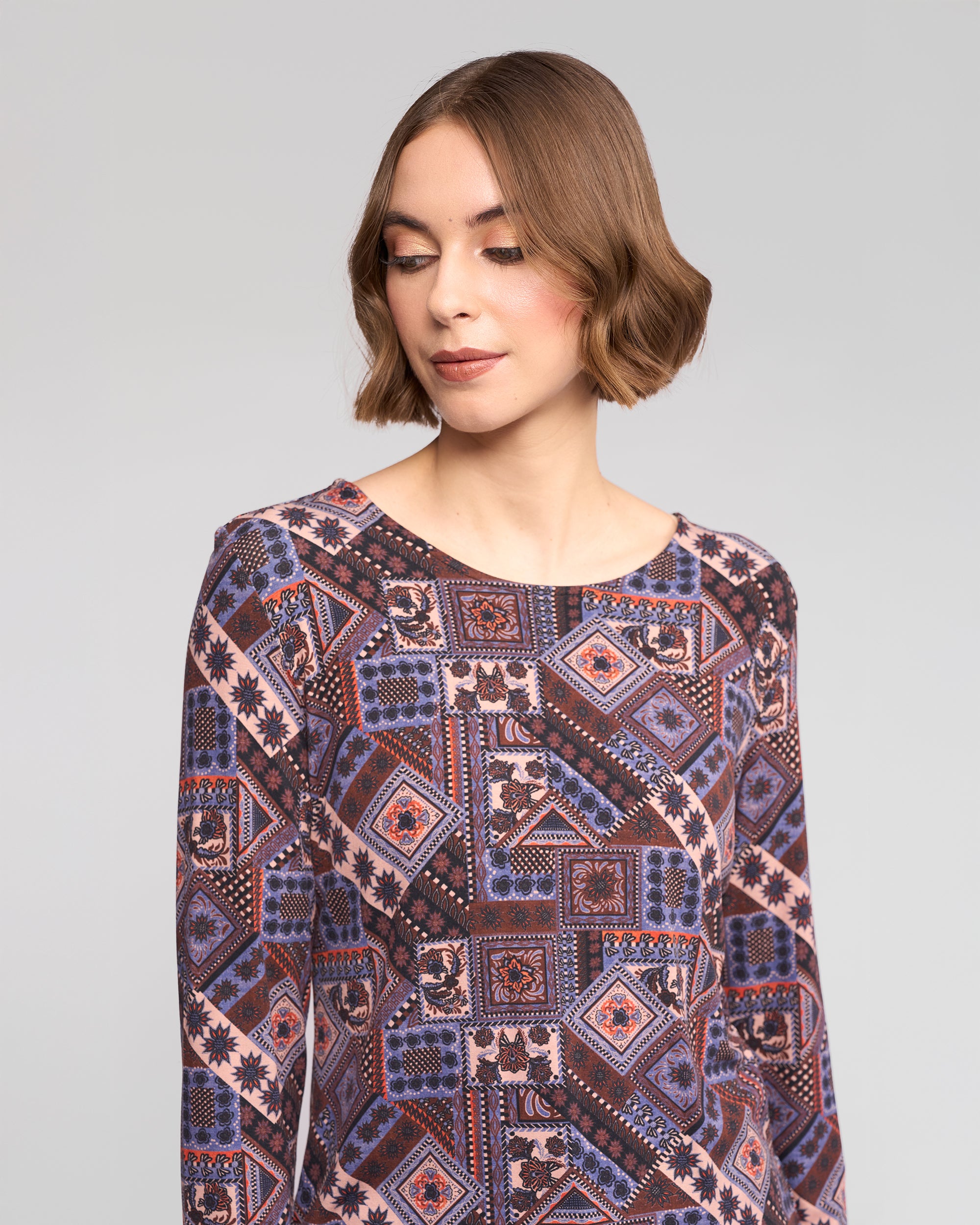 A woman with short brown hair wears the Vassalli Staple Top in Rue, showcasing its geometric multicolored print in purple, blue, and pink as she looks down and slightly to the side against a plain gray background.