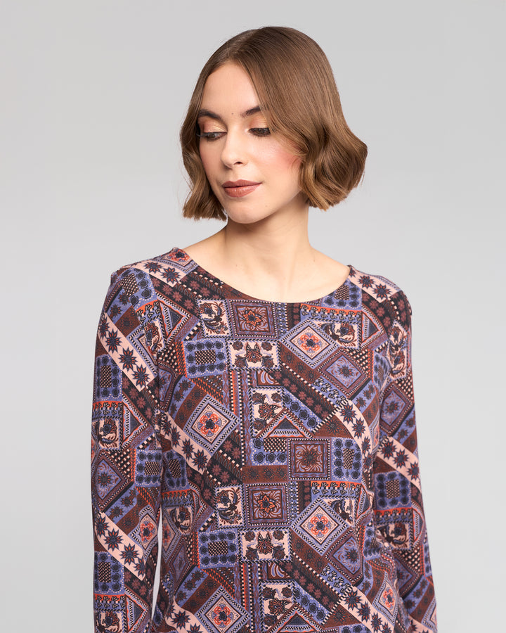 A woman with short brown hair wears the Vassalli Staple Top in Rue, showcasing its geometric multicolored print in purple, blue, and pink as she looks down and slightly to the side against a plain gray background.
