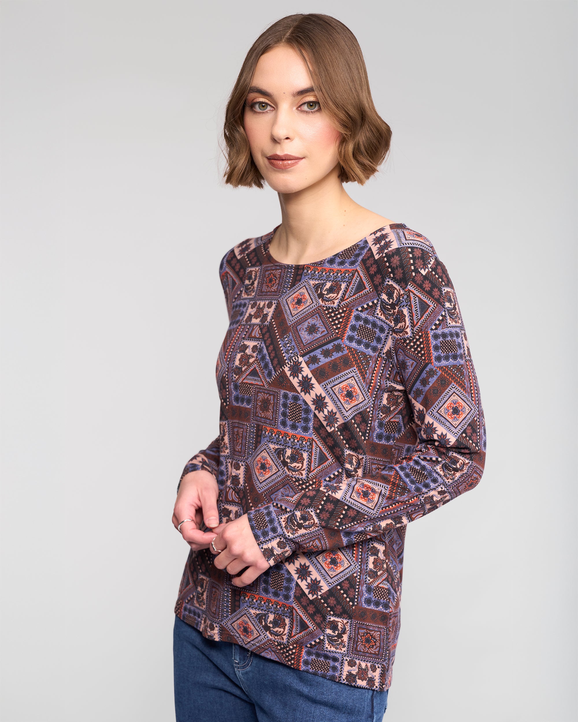 A woman with short brown hair wears the Vassalli Staple Top - Rue in stretch jersey and blue jeans, standing against a plain light gray background and looking at the camera.