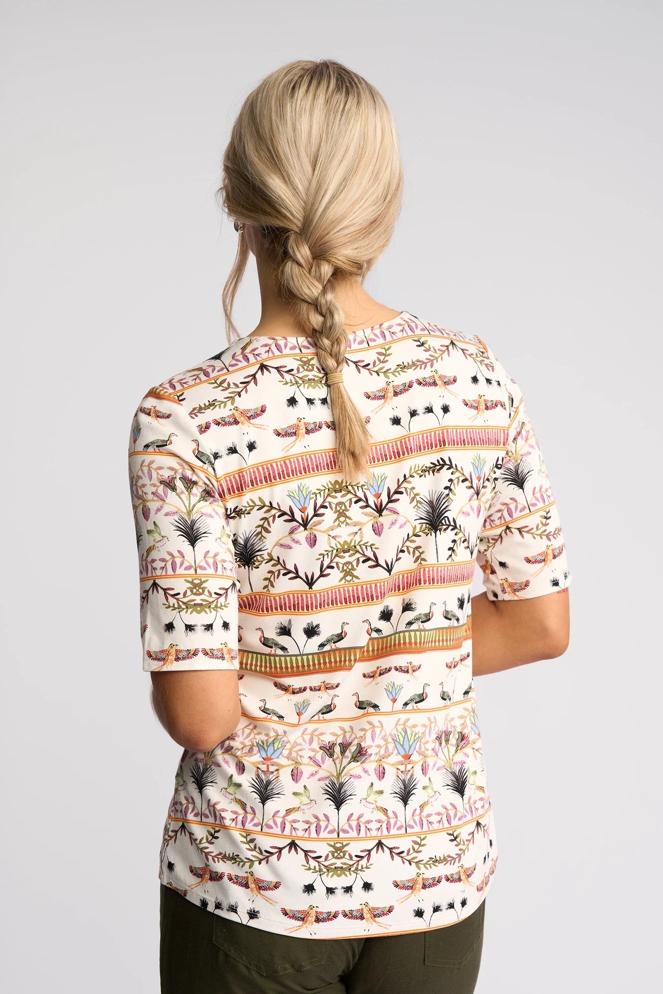 A woman with blonde hair in a braid is seen from behind, wearing the Vassalli Carmen Top - Arli, which showcases colorful, nature-inspired horizontal patterns on soft rayon-elastane fabric. She pairs it with dark green pants and stands against a plain light background.