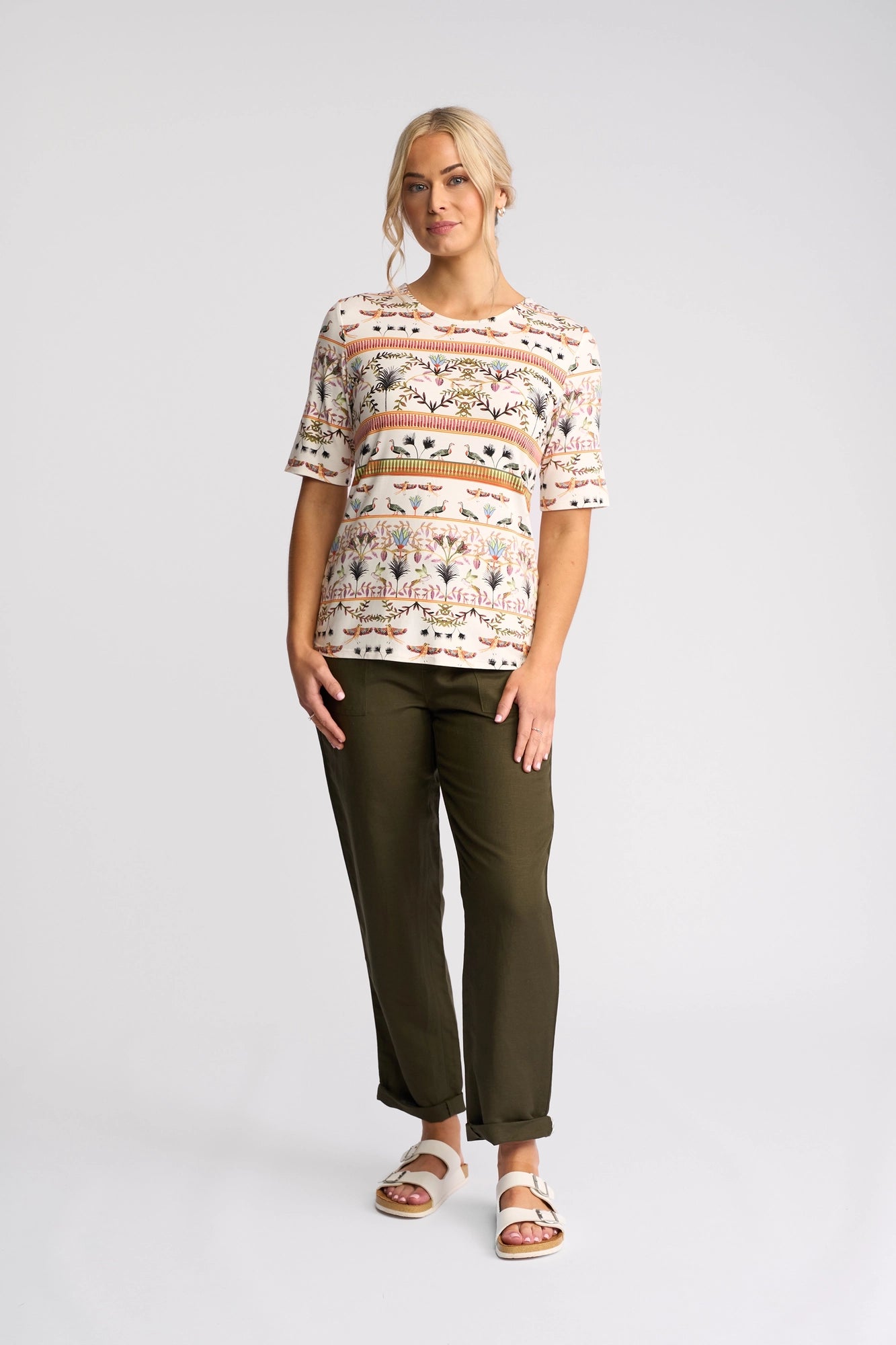 A woman poses against a simple backdrop, dressed in the Vassalli Carmen Top - Arli. The eye-catching top features a lively Arli print and short sleeves, made from comfortable rayon-elastane fabric. She pairs it with olive green pants and beige sandals, her blonde hair pulled back as she stands with one hand in her pocket.