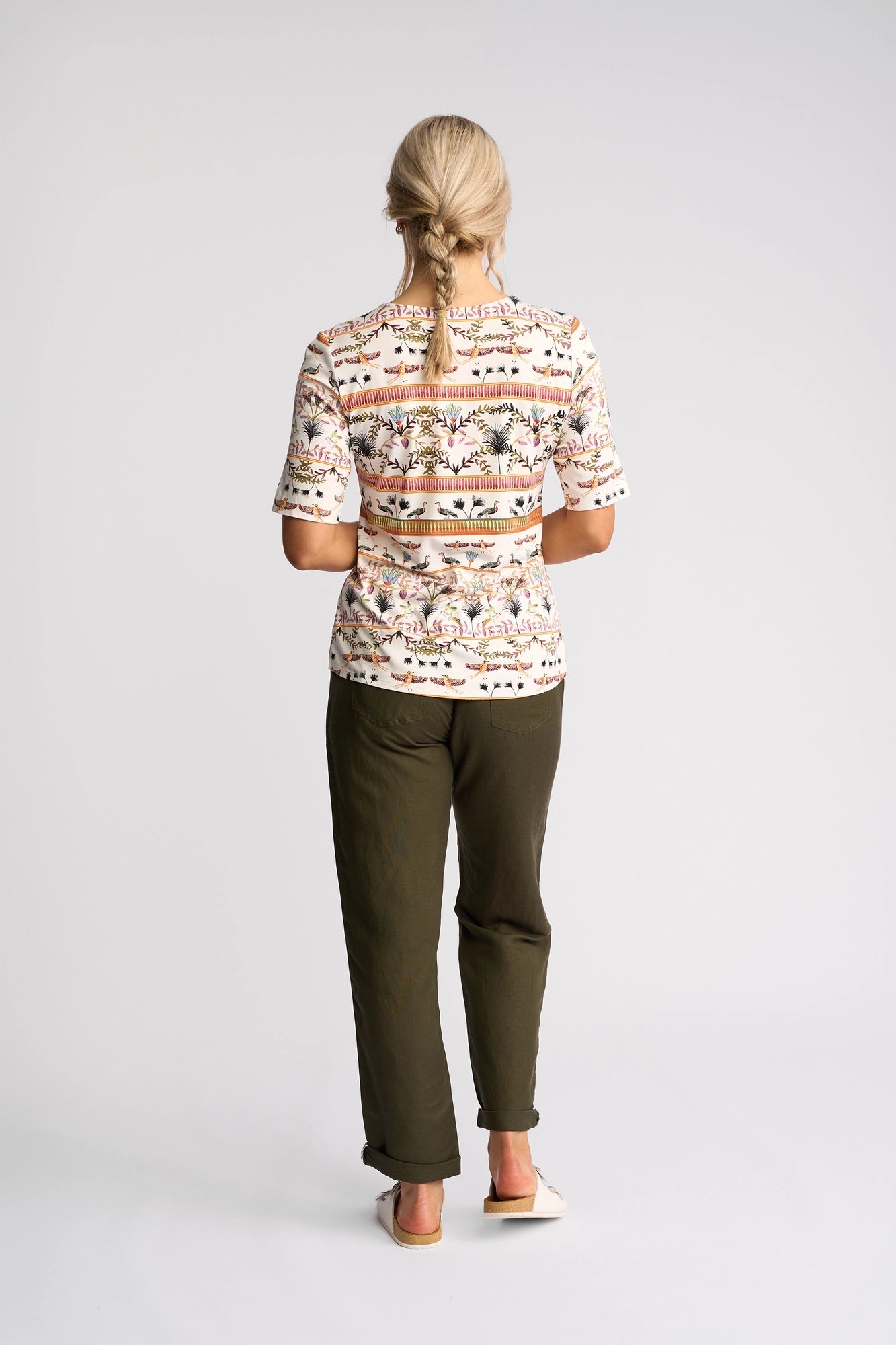 A person with blonde hair styled in a ponytail is seen from behind in a plain studio, wearing the Vassalli Carmen Top - Arli—a short-sleeve shirt in the distinctive Arli print—paired with olive green pants and white shoes.