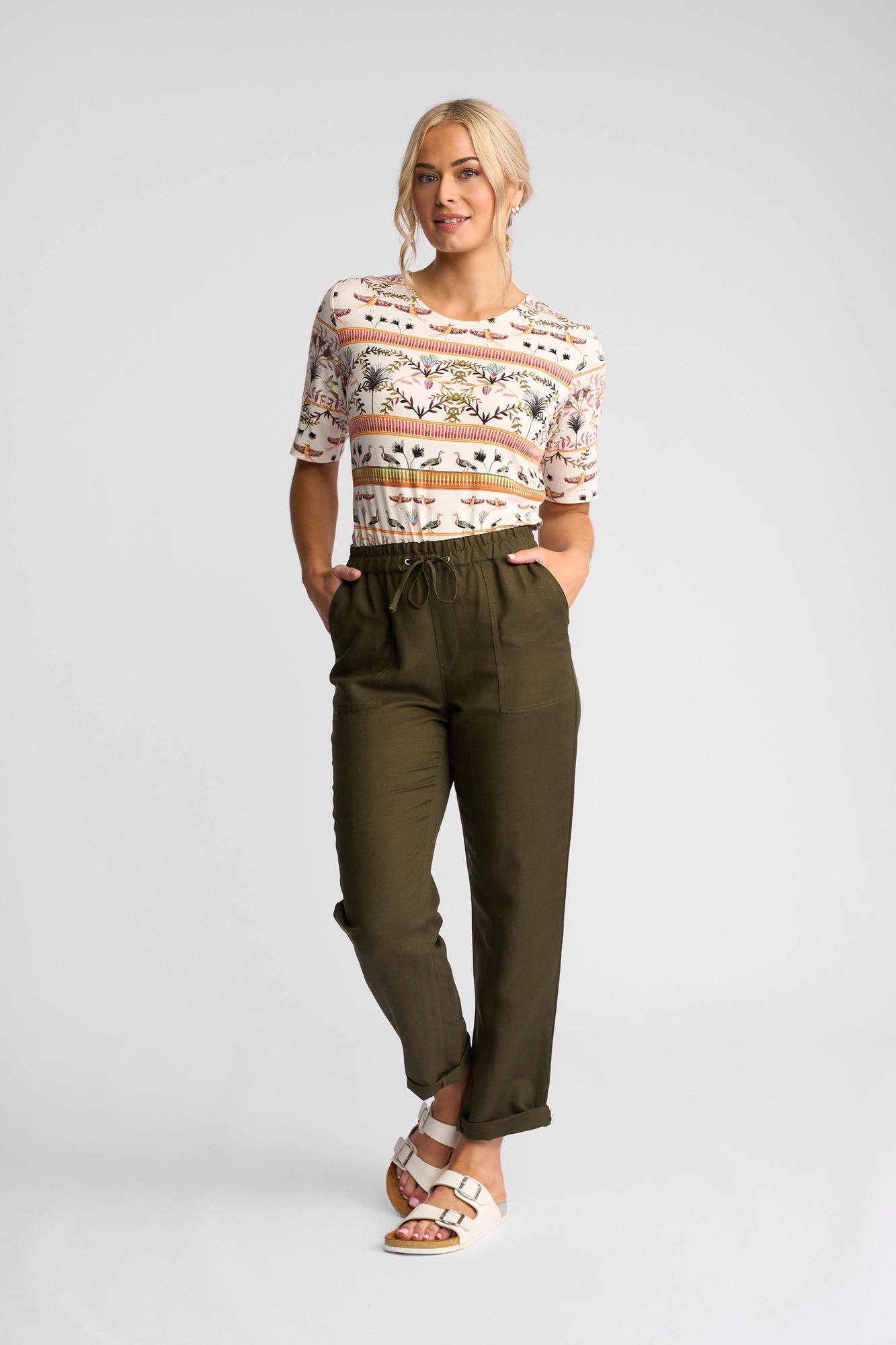 A woman stands against a plain background wearing the Vassalli Carmen Top - Arli, made from soft rayon-elastane, styled with olive green pants and white sandals. She poses with her hands in her pockets and one leg slightly bent.