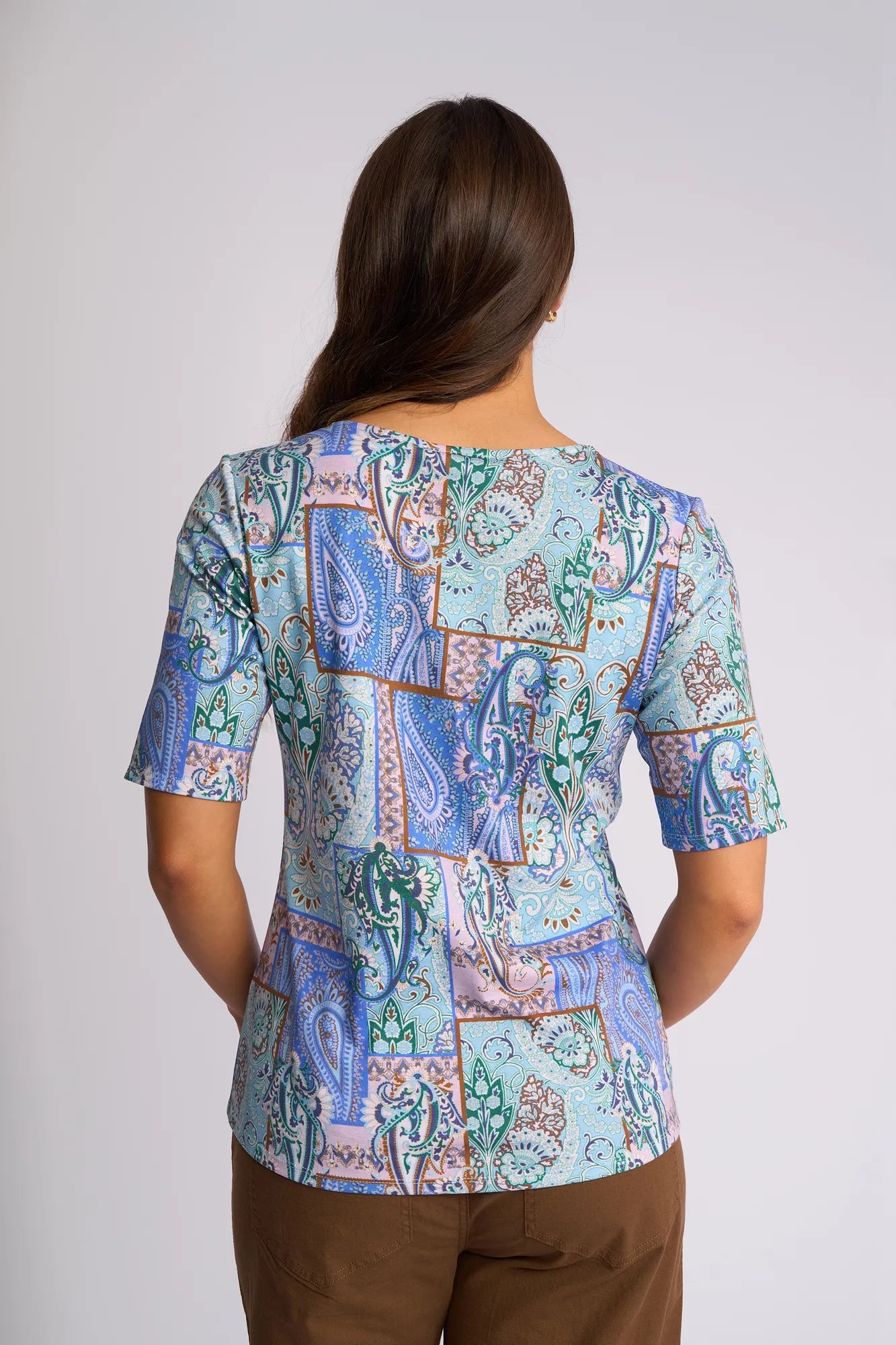 A person with long brown hair, seen from behind against a plain light background, is wearing the Vassalli Carmen Top in the Kiki print—a pastel patchwork-patterned 100% cotton shirt in blue, green, and purple tones—paired with brown pants.
