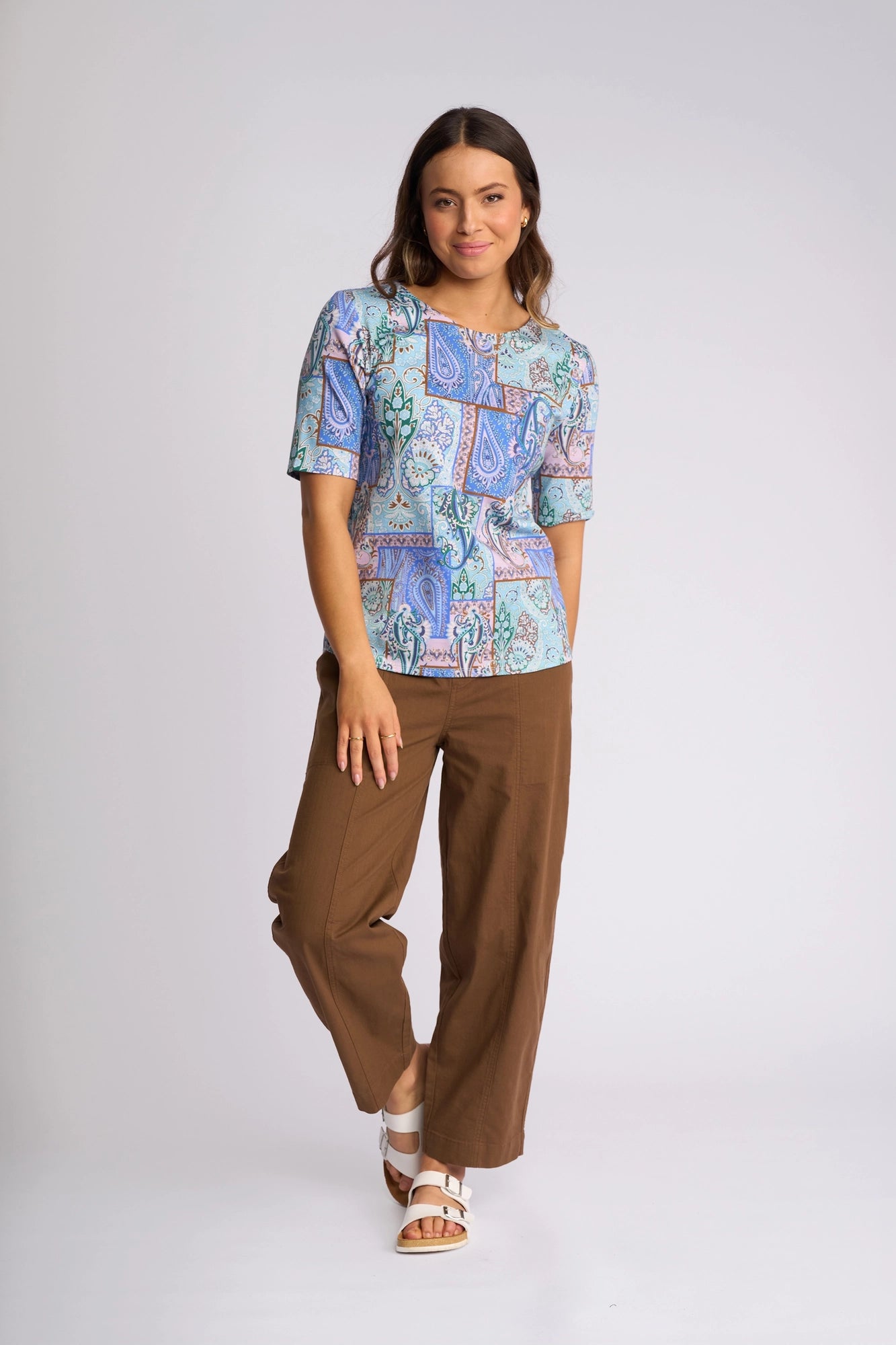 A woman models the Vassalli Carmen Top - Kiki, showcasing its vibrant Kiki print and 100% cotton fabric, paired with brown wide-leg pants and white sandals, set against a simple light background.