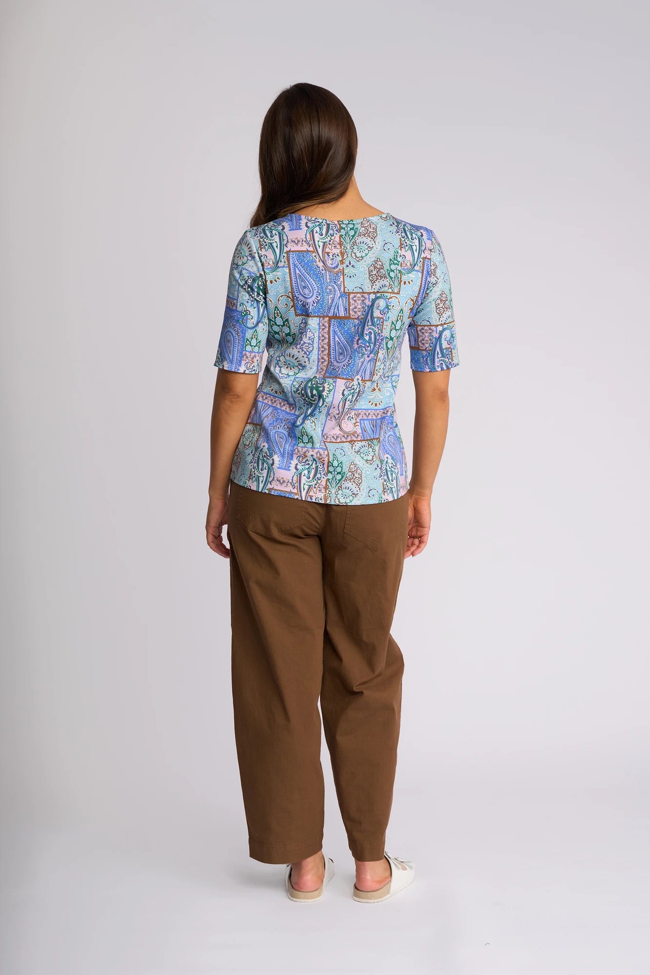 A woman stands facing away, wearing a Vassalli Carmen Top - Kiki in a blue pattern made from 100% cotton, paired with brown wide-leg pants and white shoes, against a plain light background.