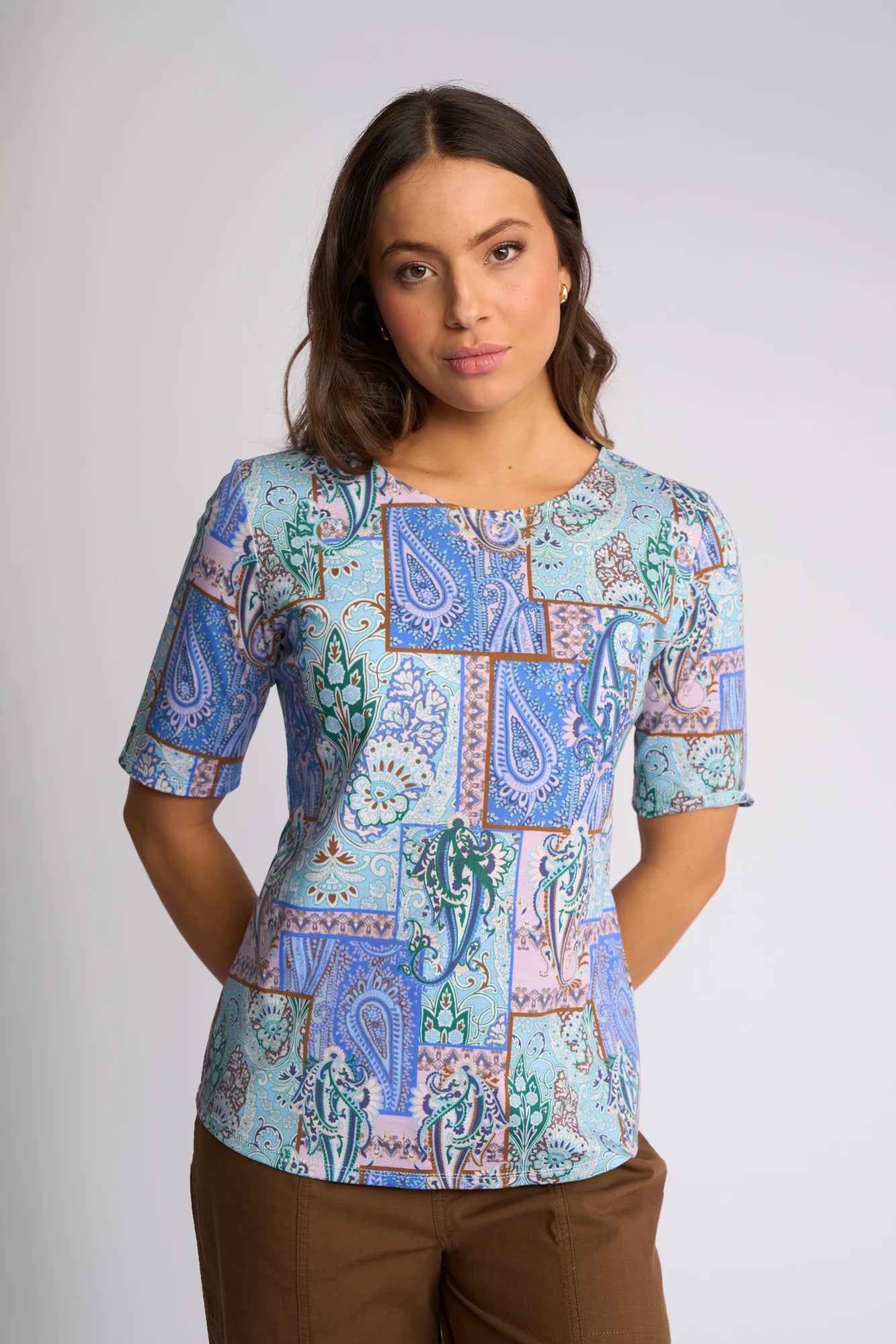 A woman with long brown hair is wearing a Vassalli Carmen Top – Kiki, crafted from 100% cotton and featuring a blue, lavender, and teal paisley print. She pairs it with brown pants and stands against a plain light background with her hands behind her back.