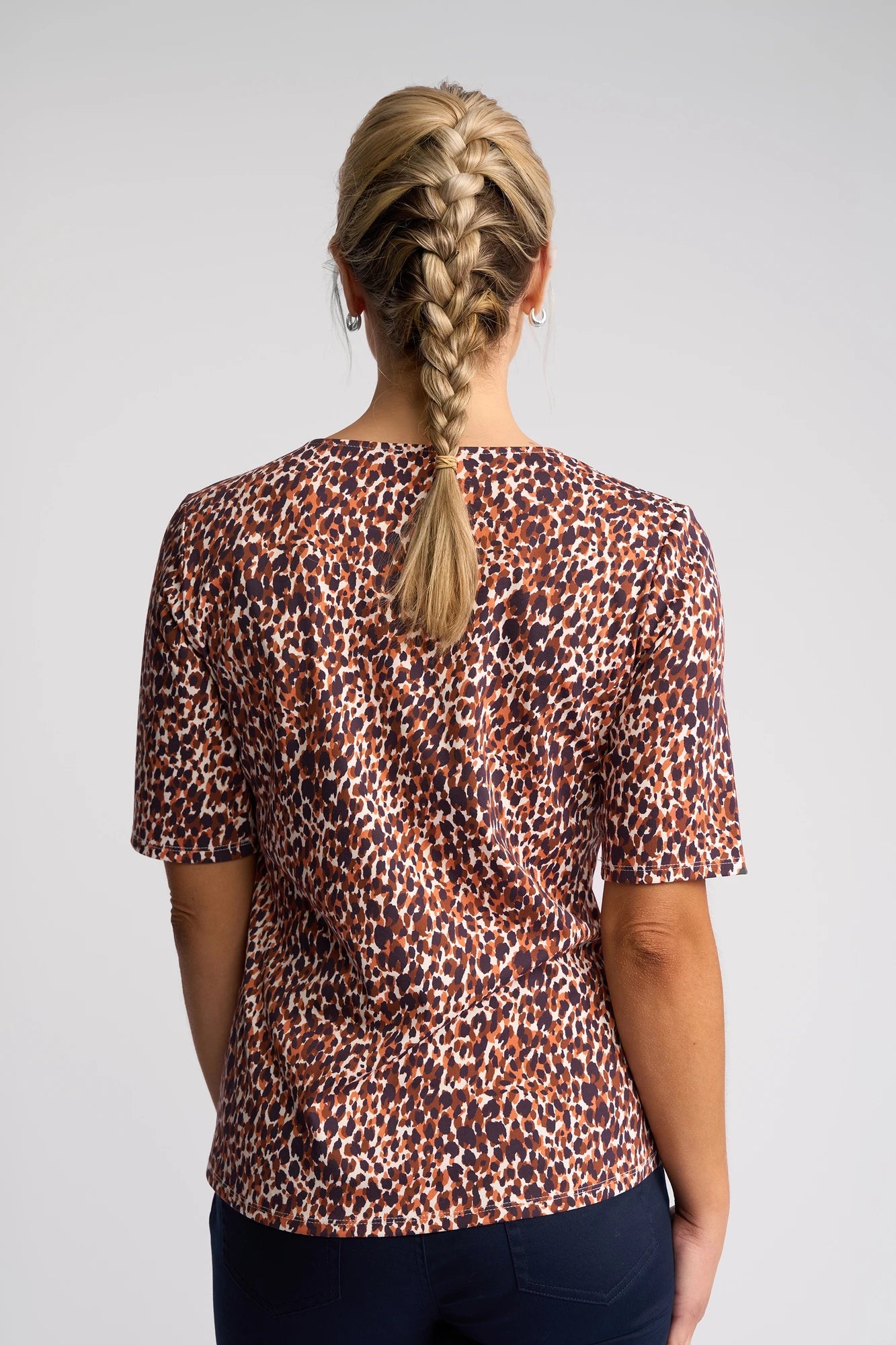 A person with long blonde hair in a braid, wearing the Vassalli Carmen Top - Zella, a short-sleeved brown and orange print blouse made from 100% cotton, paired with dark pants, stands with their back to the camera against a plain light background.