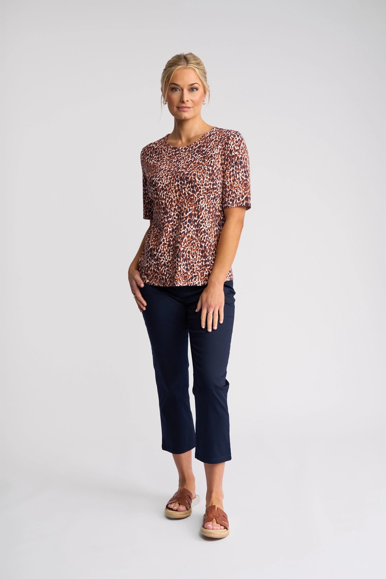 A woman with blonde hair tied back poses in front of a white background, wearing the Vassalli Carmen Top - Zella, featuring a bold print and short sleeves. She pairs the top with navy pants and brown sandals, looking at the camera in a relaxed manner. Made from 100% cotton, this top offers all-day comfort.