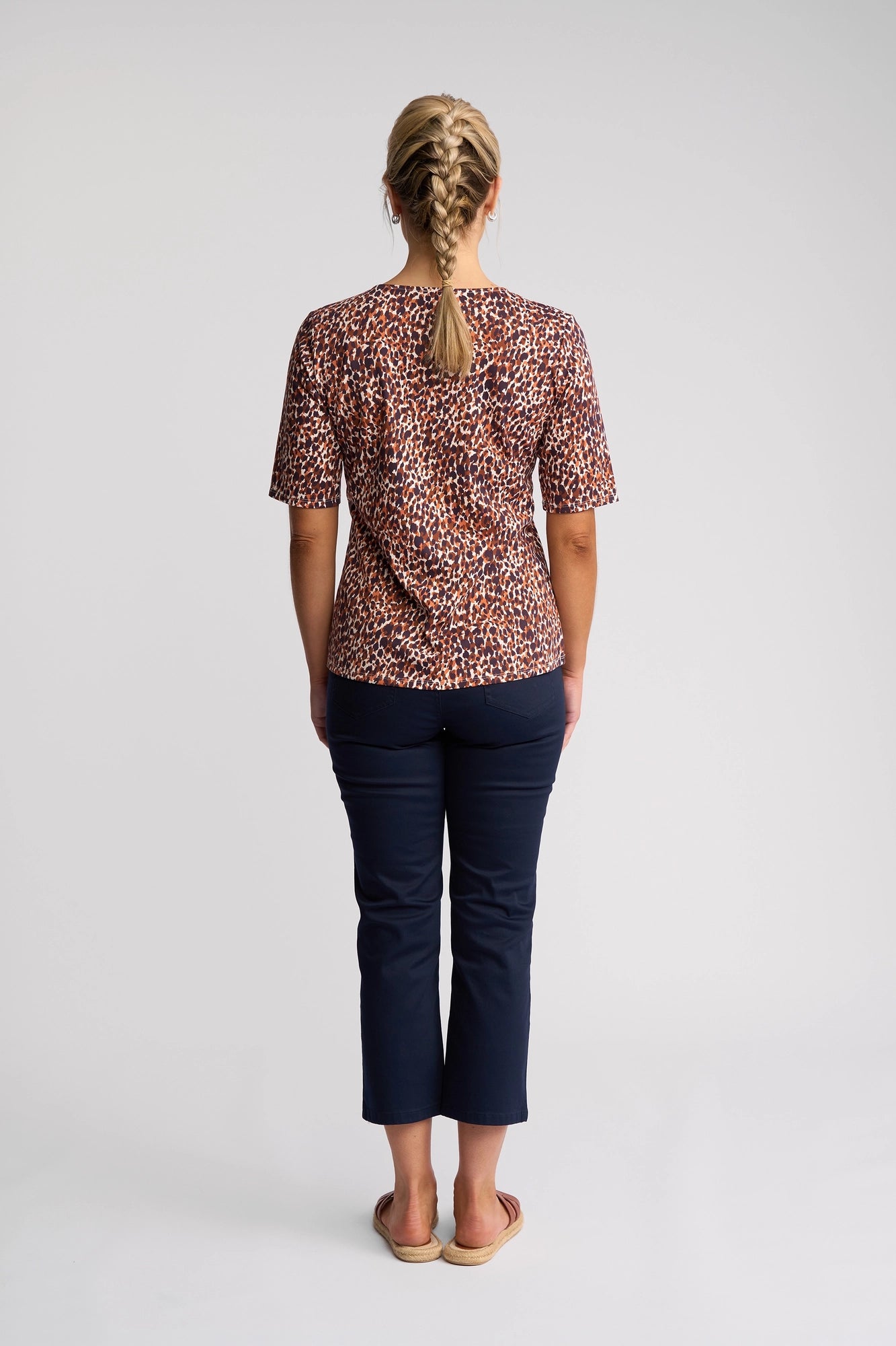 A woman with blonde hair in two braids stands facing away, wearing the Vassalli Carmen Top - Zella, a short-sleeved design crafted from 100% cotton in the striking Zella print. She pairs it with navy cropped pants and sandals, set against a plain white background.