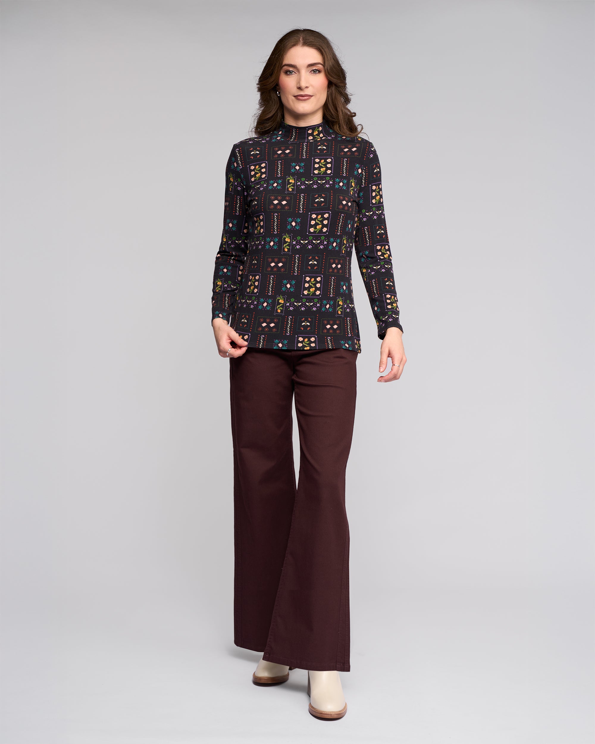 A woman poses against a plain light gray background wearing the Vassalli Foundation Top in Haven print, dark brown wide-leg pants, and cream shoes.