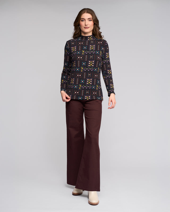 A woman poses against a plain light gray background wearing the Vassalli Foundation Top in Haven print, dark brown wide-leg pants, and cream shoes.