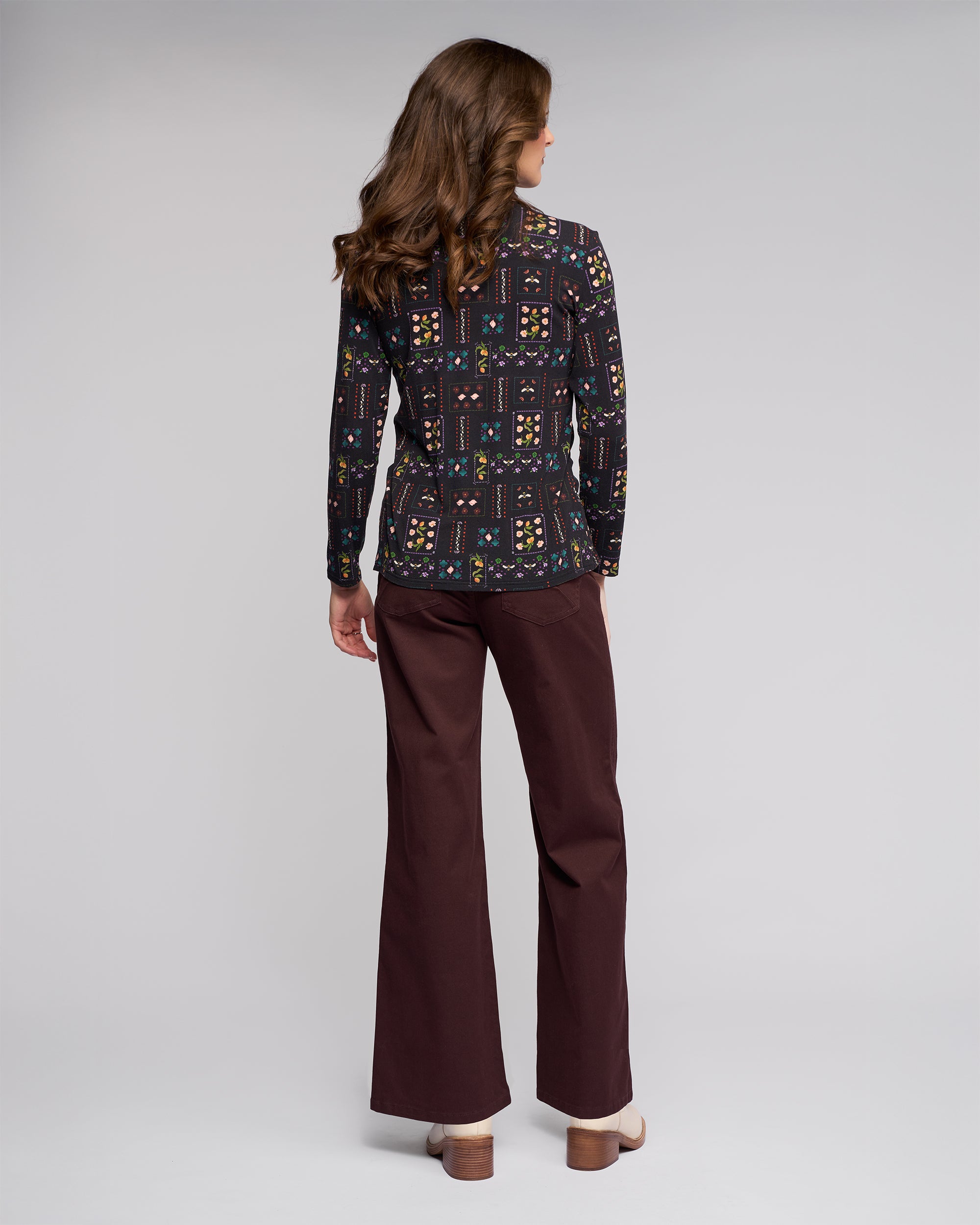 A woman with wavy brown hair faces away, wearing a black Vassalli Foundation Top - Haven and brown wide-leg pants with beige platform heels, set against a plain light gray background.