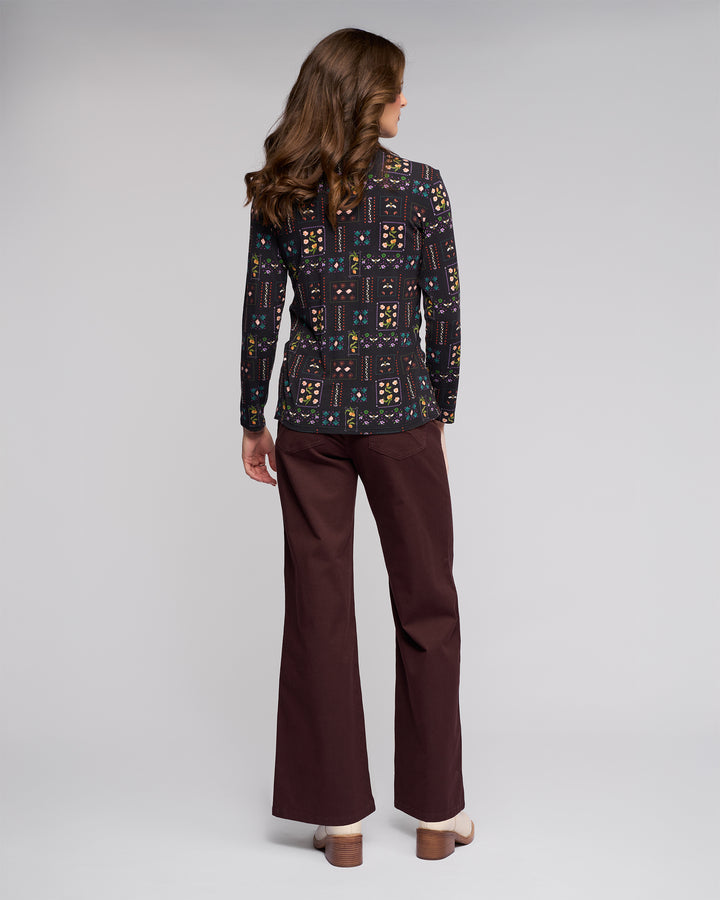 A woman with wavy brown hair faces away, wearing a black Vassalli Foundation Top - Haven and brown wide-leg pants with beige platform heels, set against a plain light gray background.