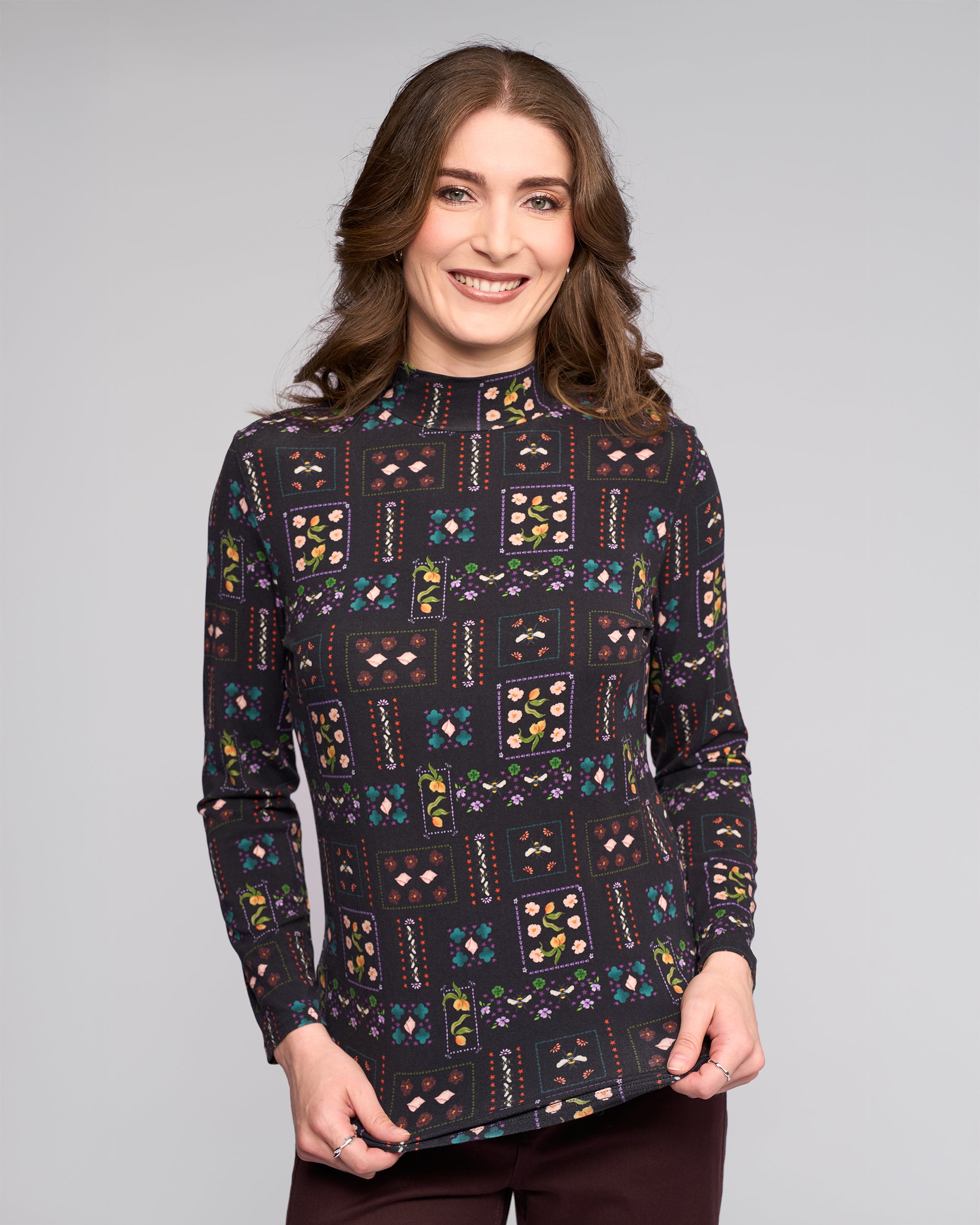 A woman with wavy brown hair smiles at the camera, wearing the Vassalli Foundation Top - Haven—a long-sleeved, high-neck stretch jersey with colorful geometric and floral patterns—standing against a plain light gray background.
