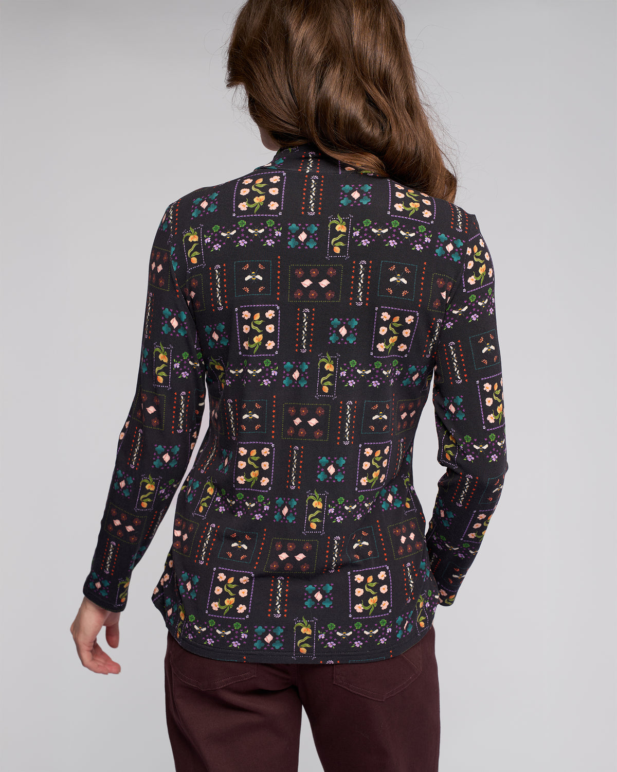 A person with long brown hair is seen from the back, wearing the Vassalli Foundation Top - Haven with a colorful patchwork print and dark maroon pants, against a plain light background.