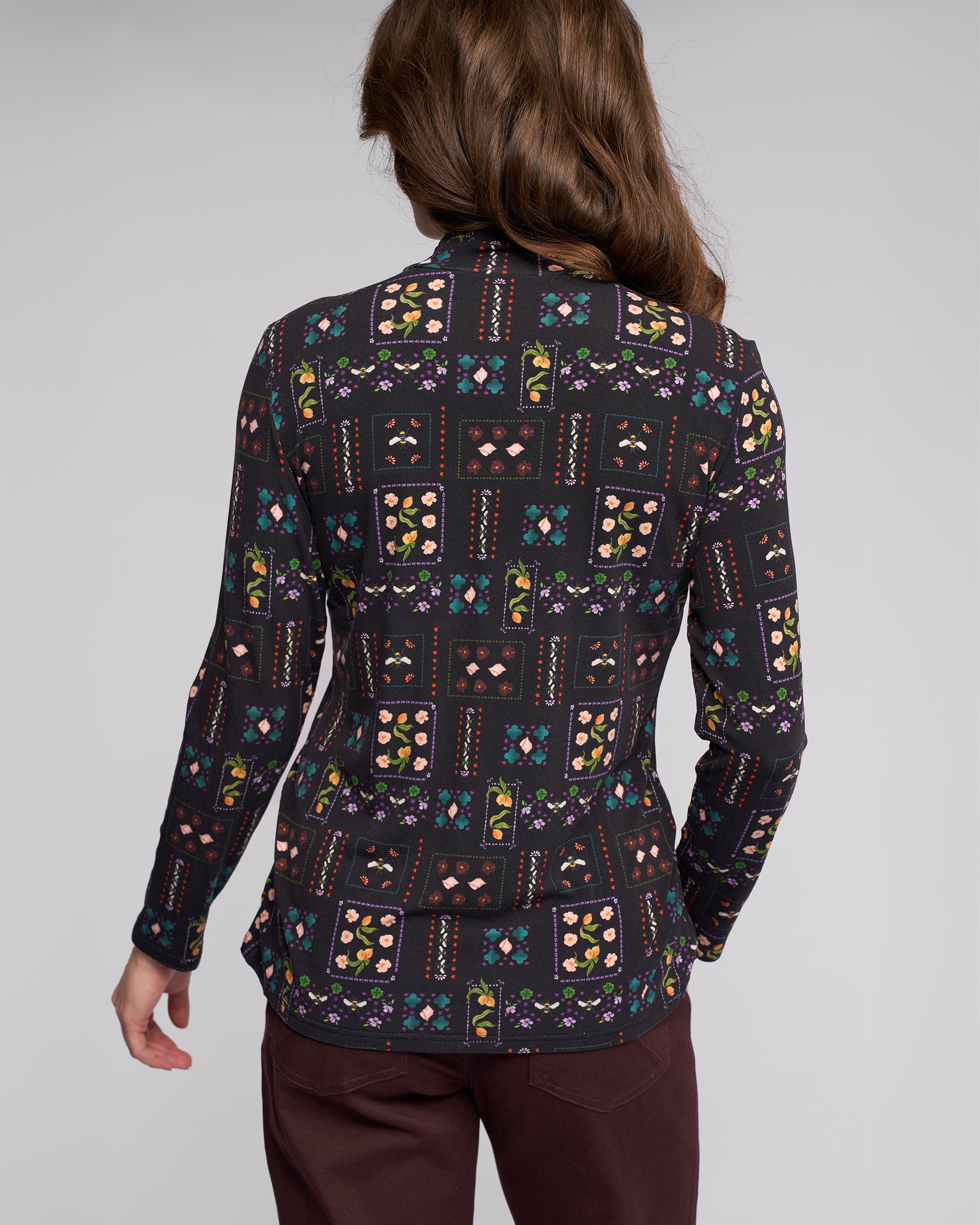 A person with long brown hair is seen from the back, wearing the Vassalli Foundation Top - Haven with a colorful patchwork print and dark maroon pants, against a plain light background.