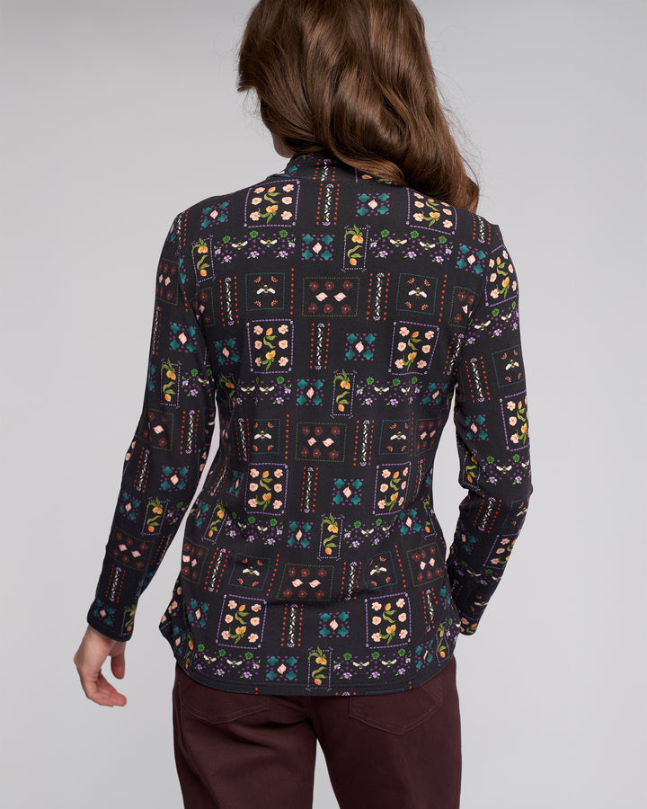 A person with long brown hair is seen from the back, wearing the Vassalli Foundation Top - Haven with a colorful patchwork print and dark maroon pants, against a plain light background.
