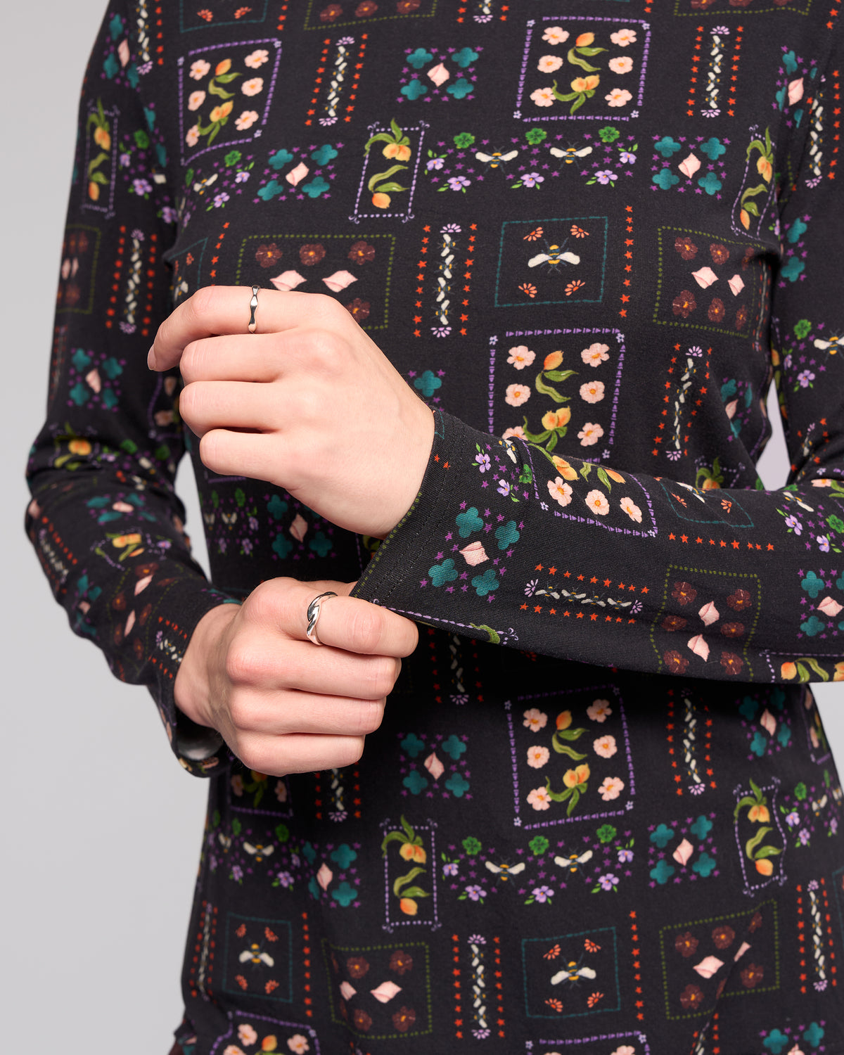 A person wears the Vassalli Foundation Top in the vibrant Haven floral patchwork, featuring long sleeves and a high neck. They hold one sleeve with their other hand, displaying rings, against a plain light background.