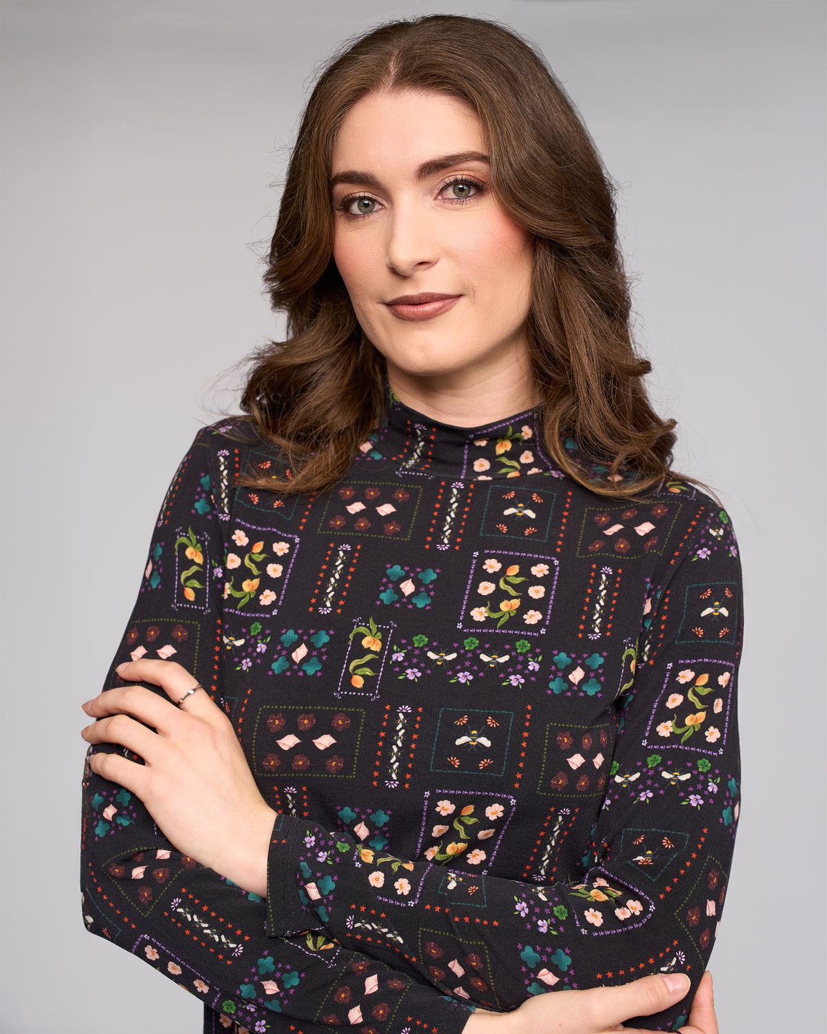 A woman with wavy brown hair and light makeup stands with arms crossed, wearing the Vassalli Foundation Top - Haven, a dark stretch jersey high-neck top featuring colorful floral and geometric prints, against a plain gray background.