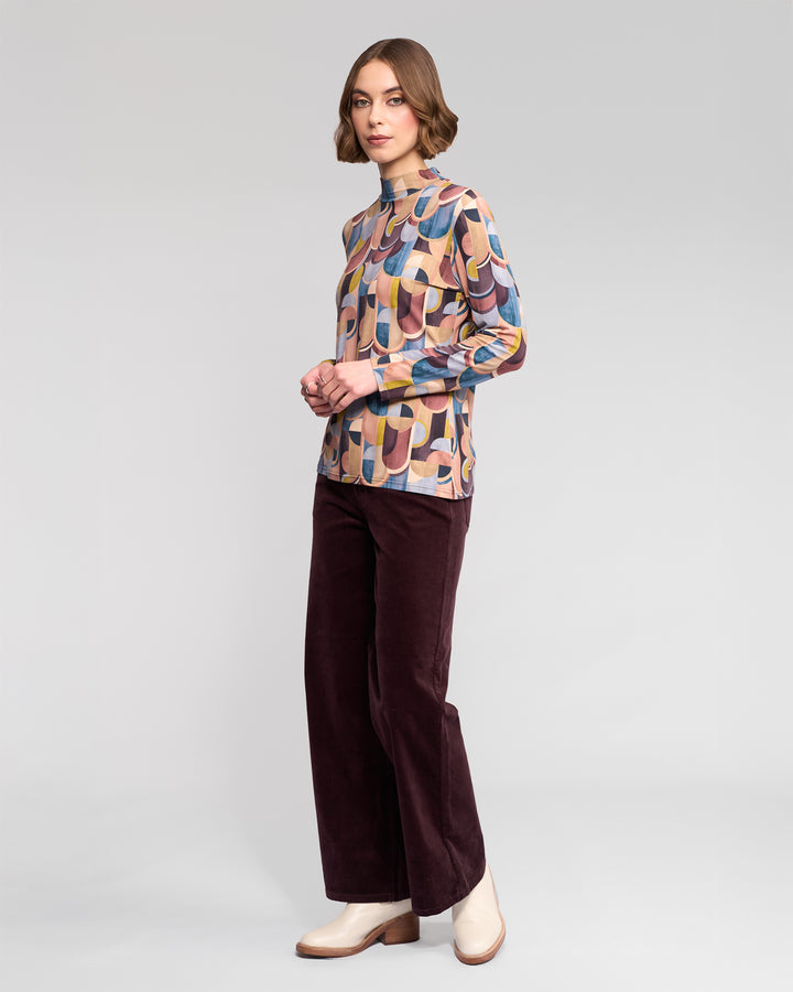 A woman with short, wavy brown hair and a neutral expression poses against a light gray background, wearing the Vassalli Foundation Top - Revolve, dark brown wide-leg pants, and cream ankle boots.