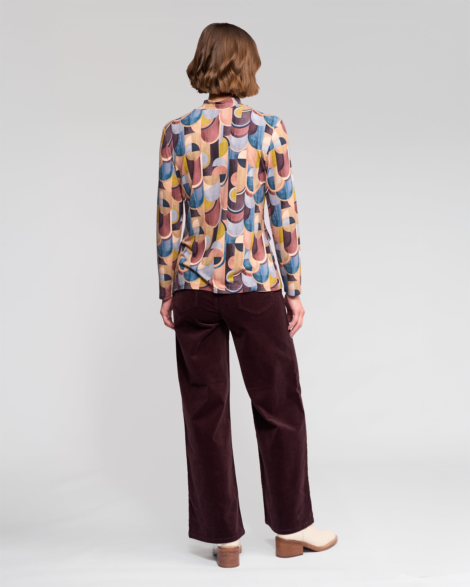A person stands facing away, wearing the Vassalli Foundation Top - Revolve with a multicolored geometric retro print, paired with dark wide-leg pants and light heeled shoes against a plain light background.