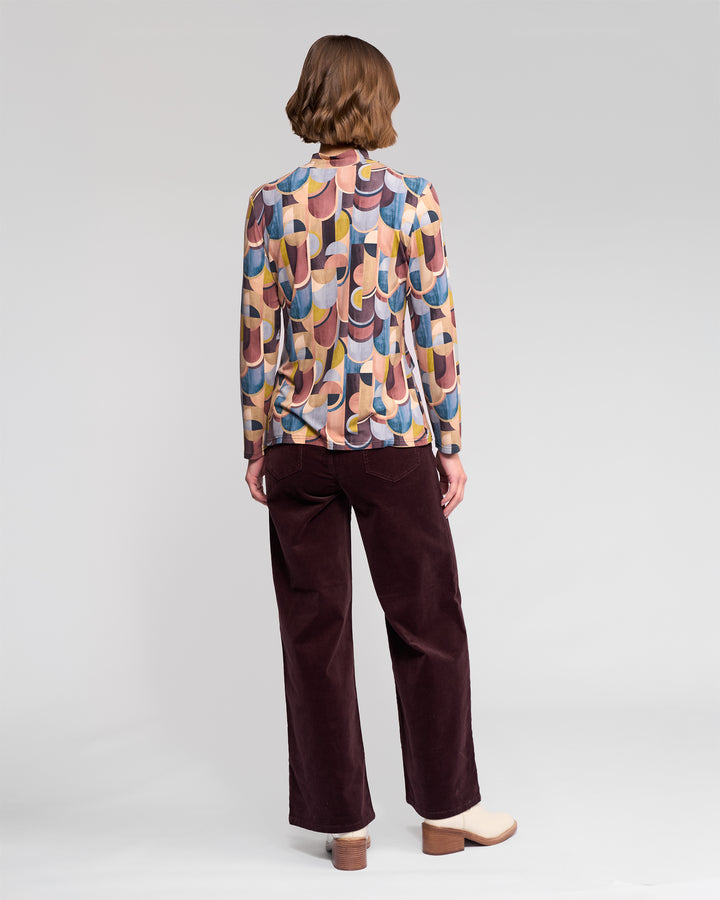 A person stands facing away, wearing the Vassalli Foundation Top - Revolve with a multicolored geometric retro print, paired with dark wide-leg pants and light heeled shoes against a plain light background.