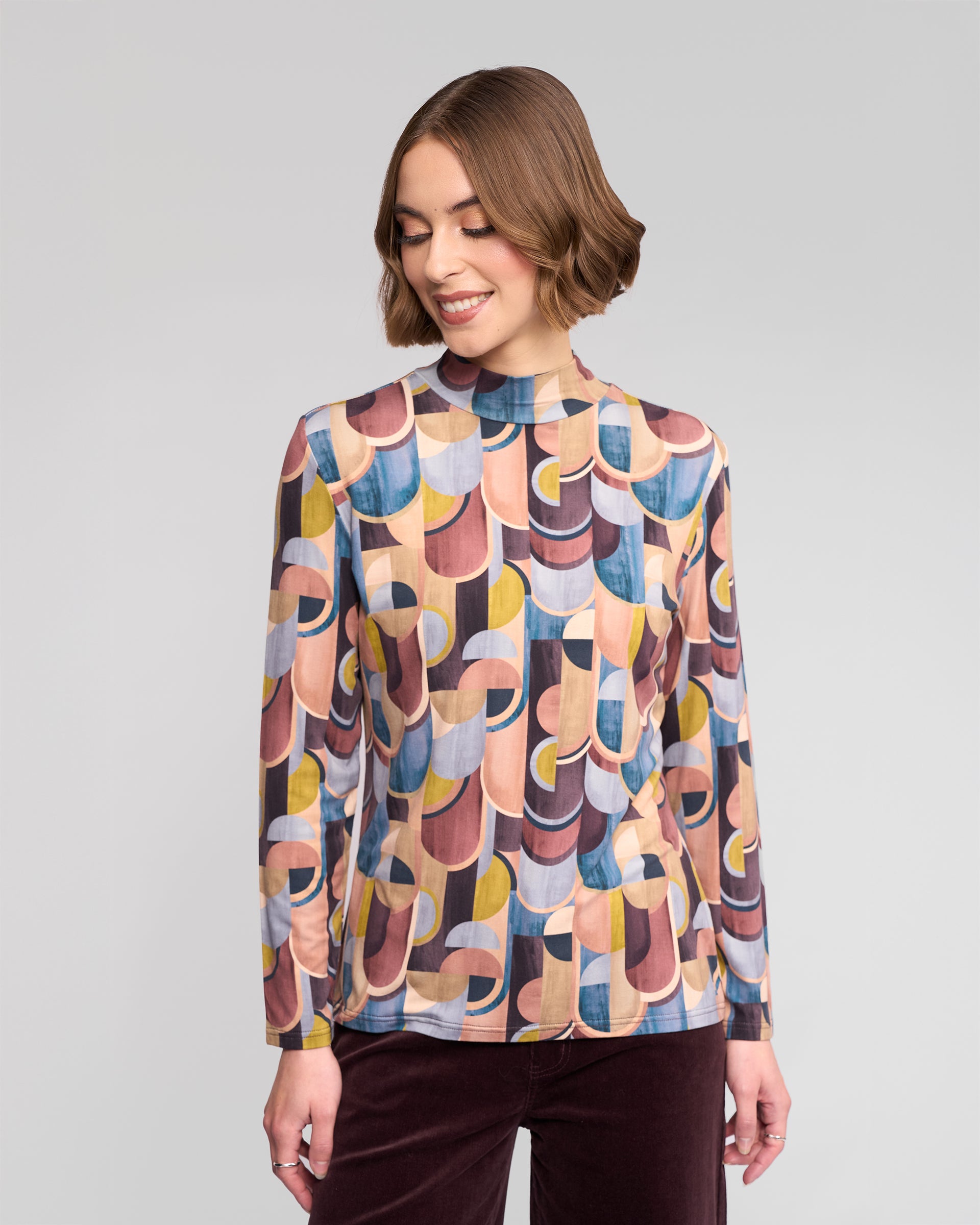 A woman with short brown hair wears the Vassalli Foundation Top - Revolve, featuring a colorful geometric pattern, paired with burgundy pants. She smiles and looks down to her right against a plain light gray background.