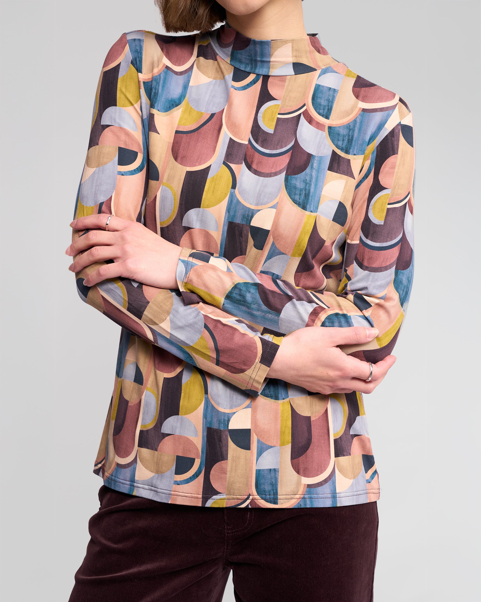 A person wears the Vassalli Foundation Top – Revolve, a long-sleeve, high-neck stretch jersey with bold geometric patterns in yellow, blue, pink, and brown, paired with dark trousers and posed against a neutral background.