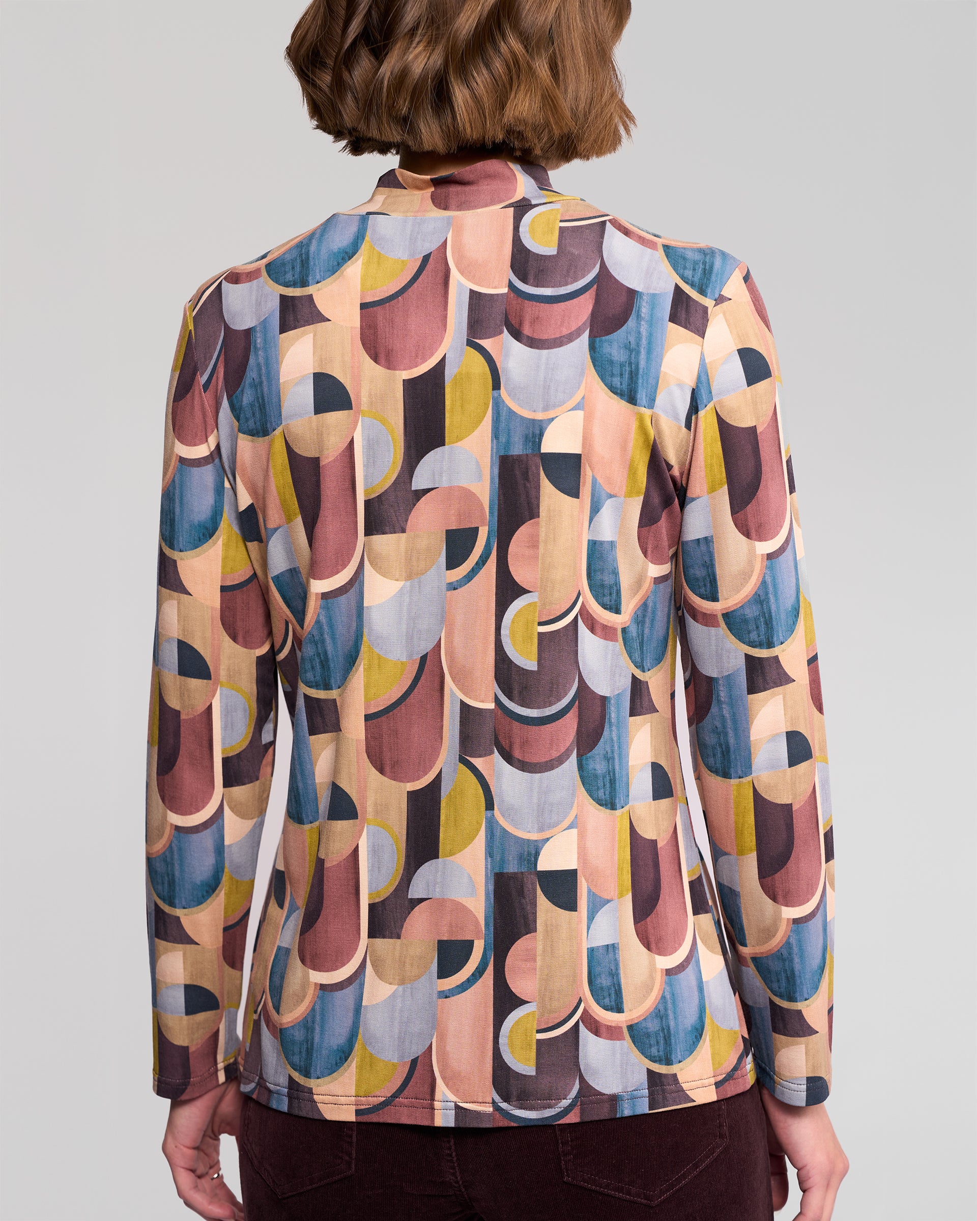A person with short brown hair is seen from behind wearing the Vassalli Foundation Top – Revolve, a long-sleeve, high-neck stretch jersey featuring retro geometric prints in pink, yellow, blue, and brown.