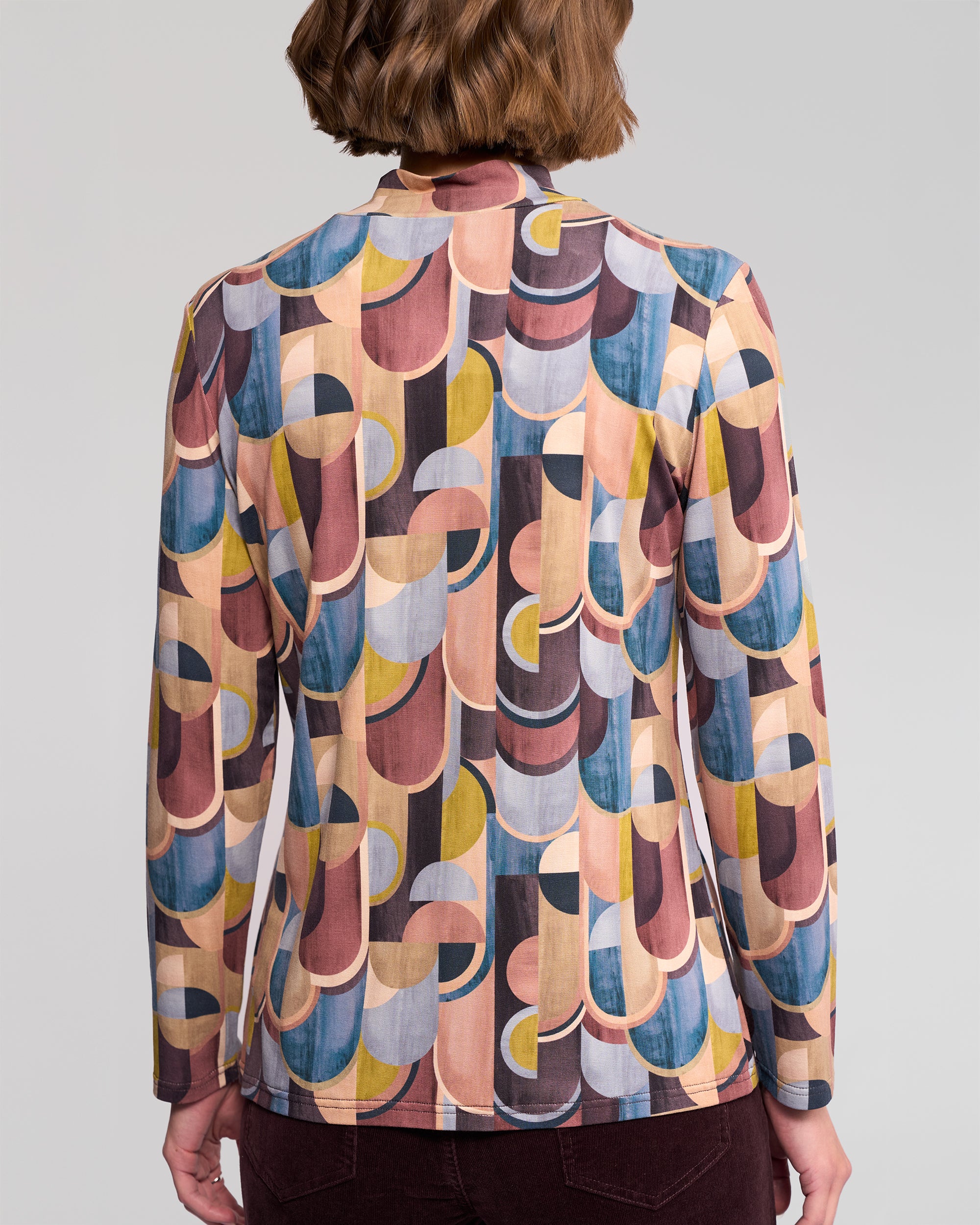 A person with short brown hair is seen from behind wearing the Vassalli Foundation Top – Revolve, a long-sleeve, high-neck stretch jersey featuring retro geometric prints in pink, yellow, blue, and brown.