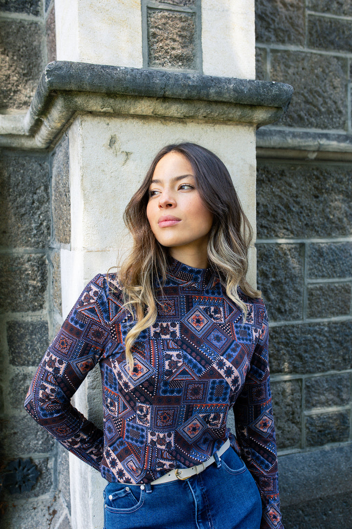 A young woman with long, wavy hair wears the Vassalli Foundation Top - Rue and high-waisted jeans as she stands in front of a stone wall, looking thoughtfully to the side.