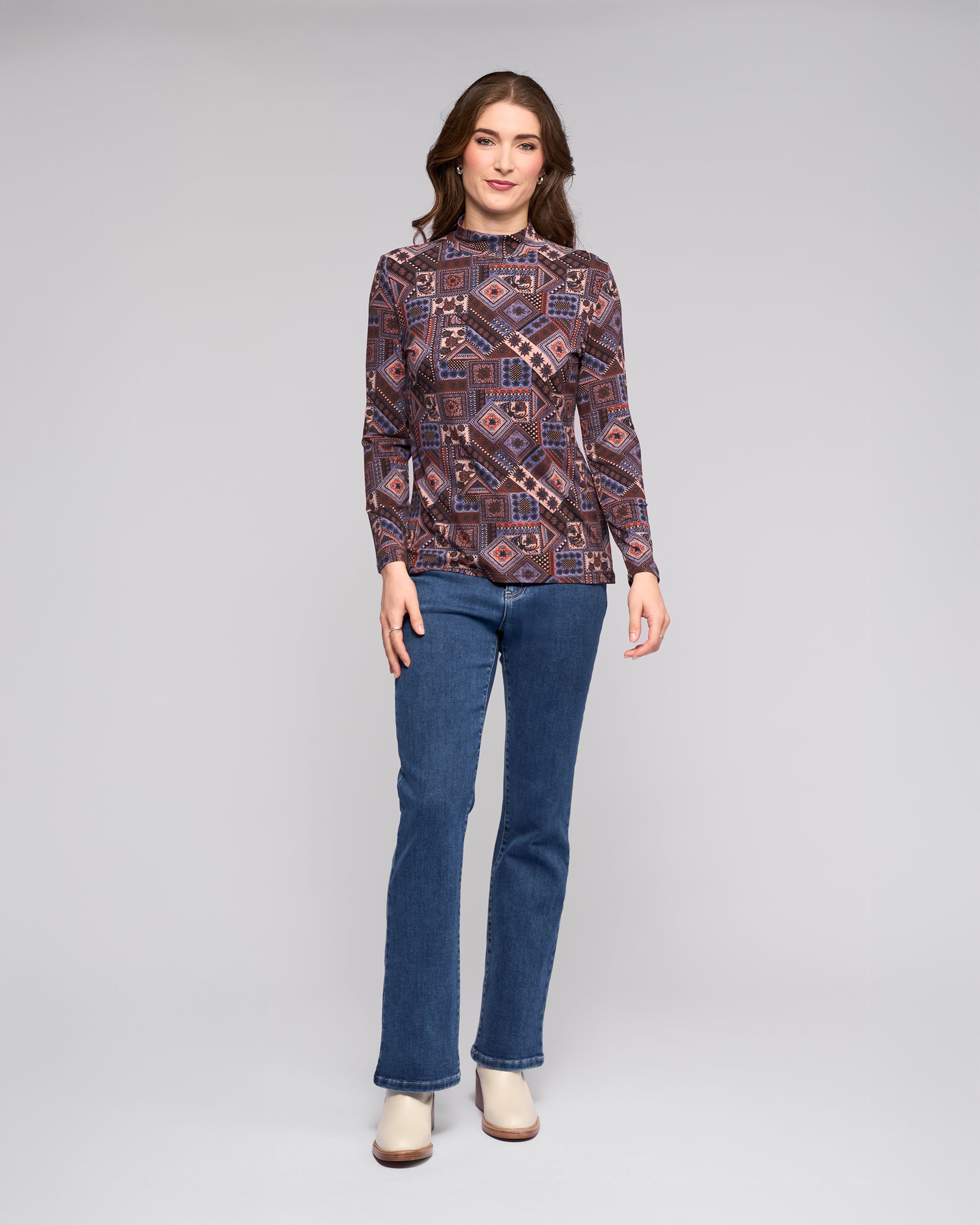A woman poses in a studio wearing the Vassalli Foundation Top - Rue, styled with blue jeans and beige ankle boots, set against a plain light gray background.